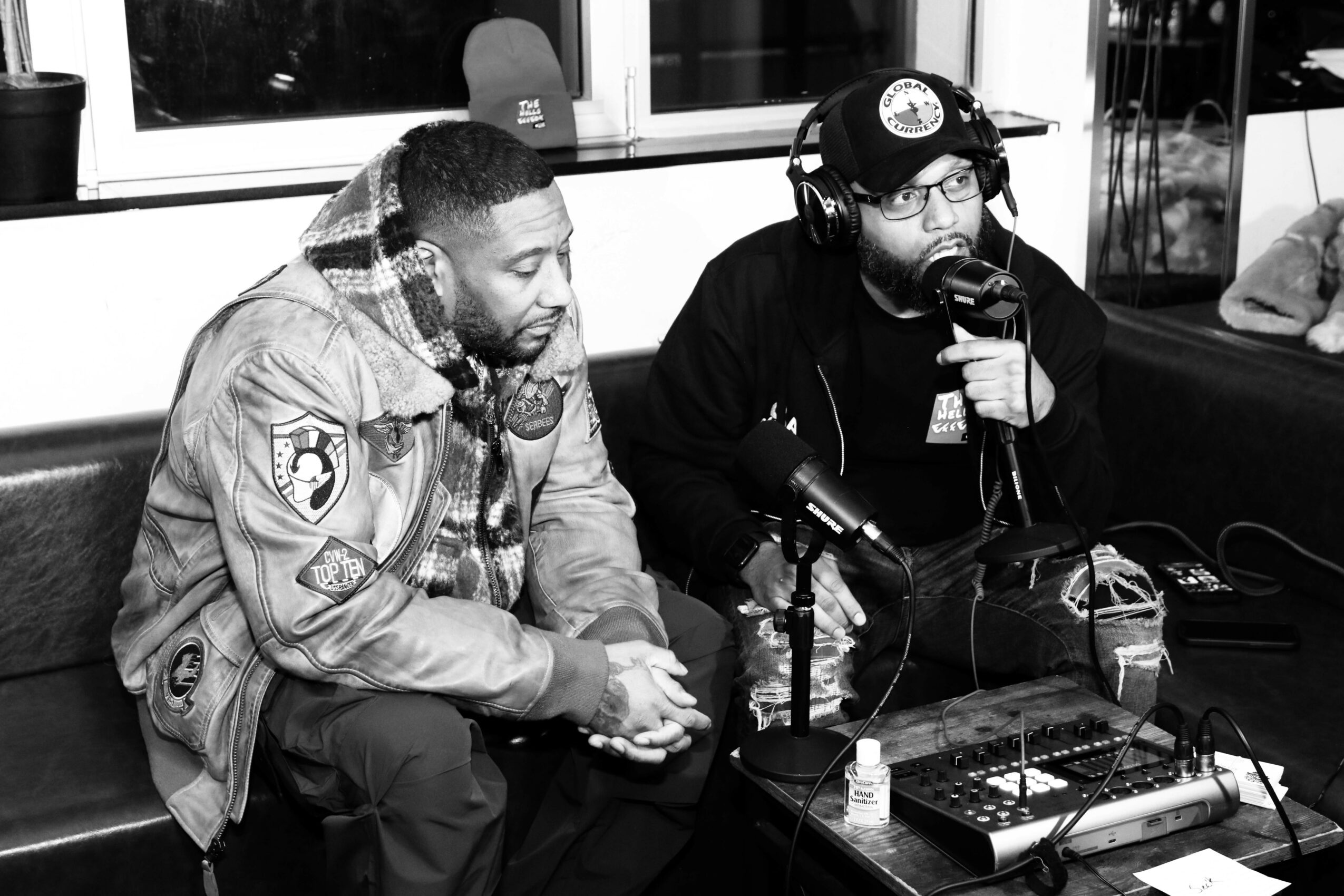 Two men sit at a table with microphones and audio equipment, one wearing a cap and headphones, the other in a jacket.