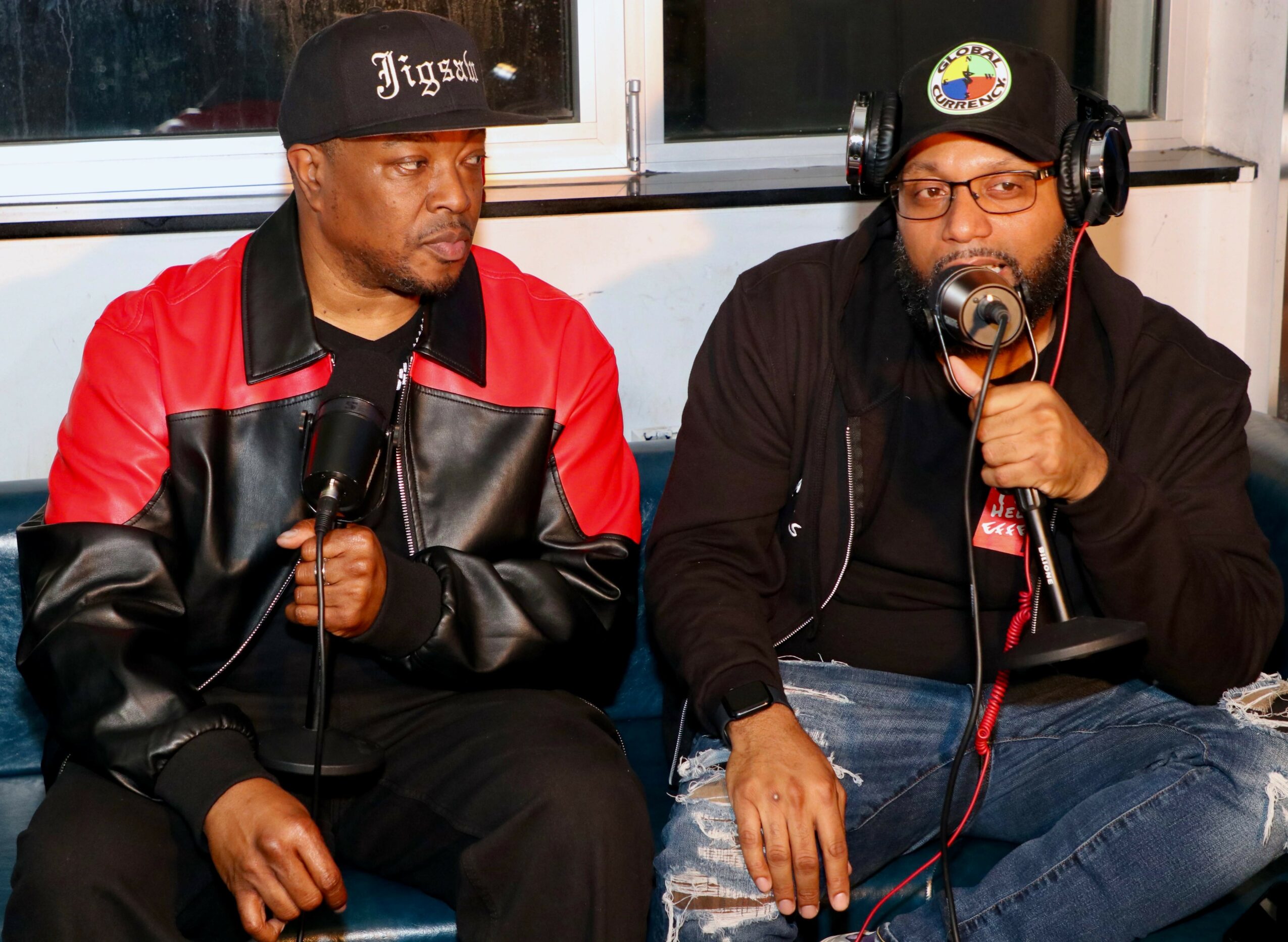 Two men sit side by side, one holding a microphone, in a room with a window behind them.
