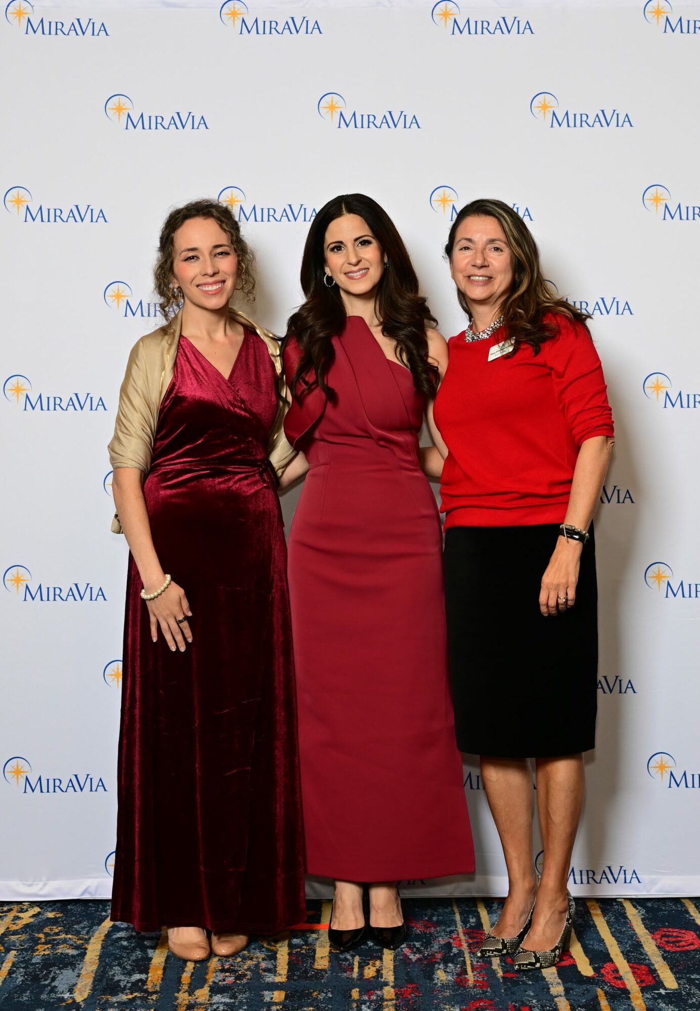 Three women standing together in front of a Miravia backdrop, smiling, dressed in elegant red and black outfits.
