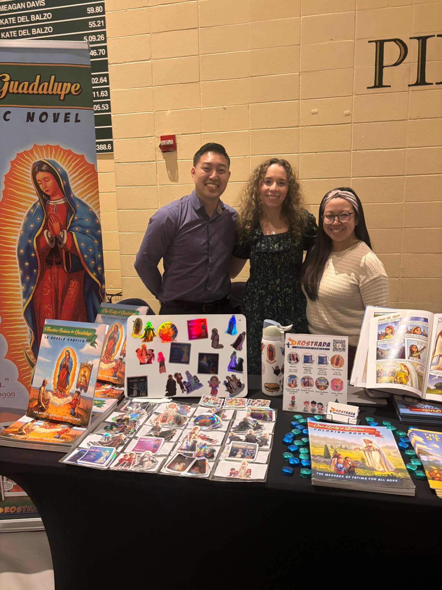 Three people standing behind a table with colorful books and merchandise, in a room with beige walls and a black tablecloth.
