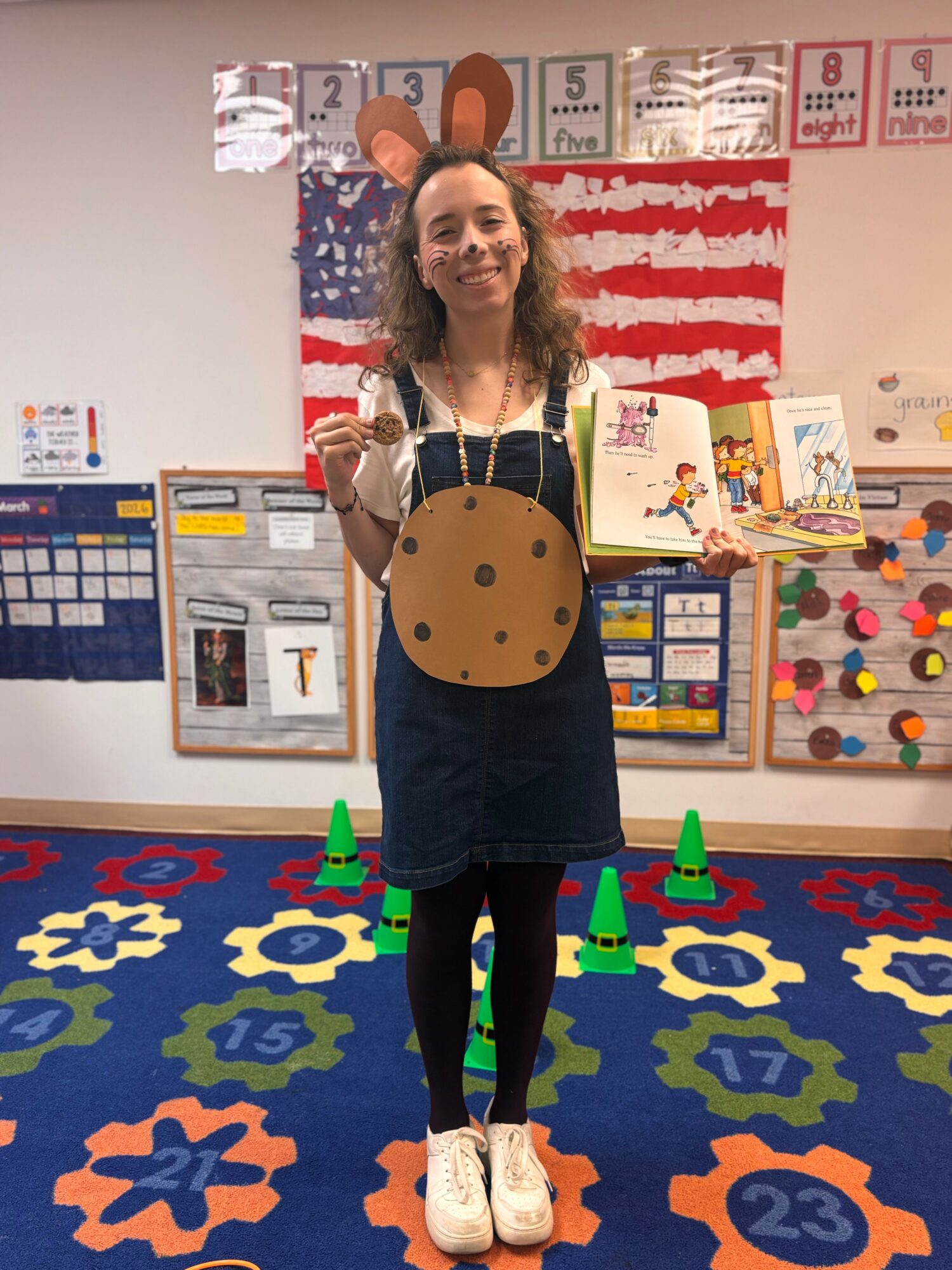 Woman standing in a colorful classroom holding a book, wearing a large round bag, with educational posters behind her.
