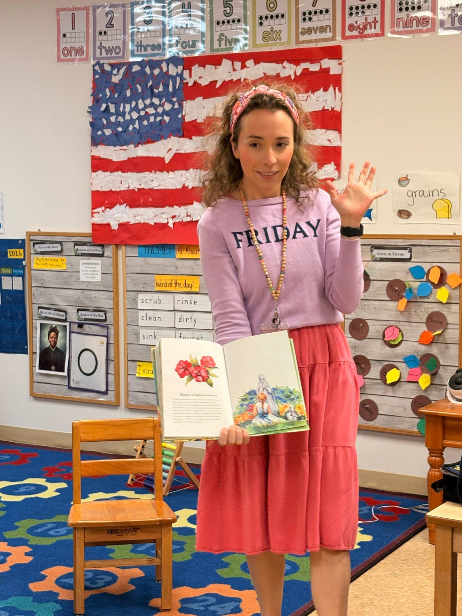 Woman standing in classroom holding open book, wearing pink skirt and purple top, with American flag display behind her.
