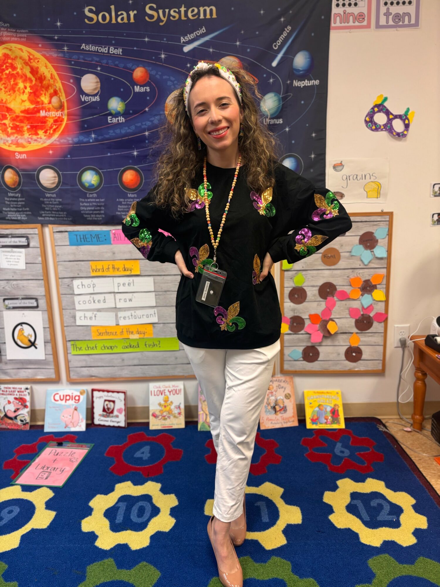 Young woman with curly hair smiling, standing in classroom with educational posters and colorful decorations.
