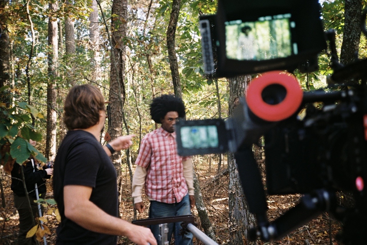 Two people in a forest, one with curly hair and a pink checkered shirt, the other with short hair, talking.