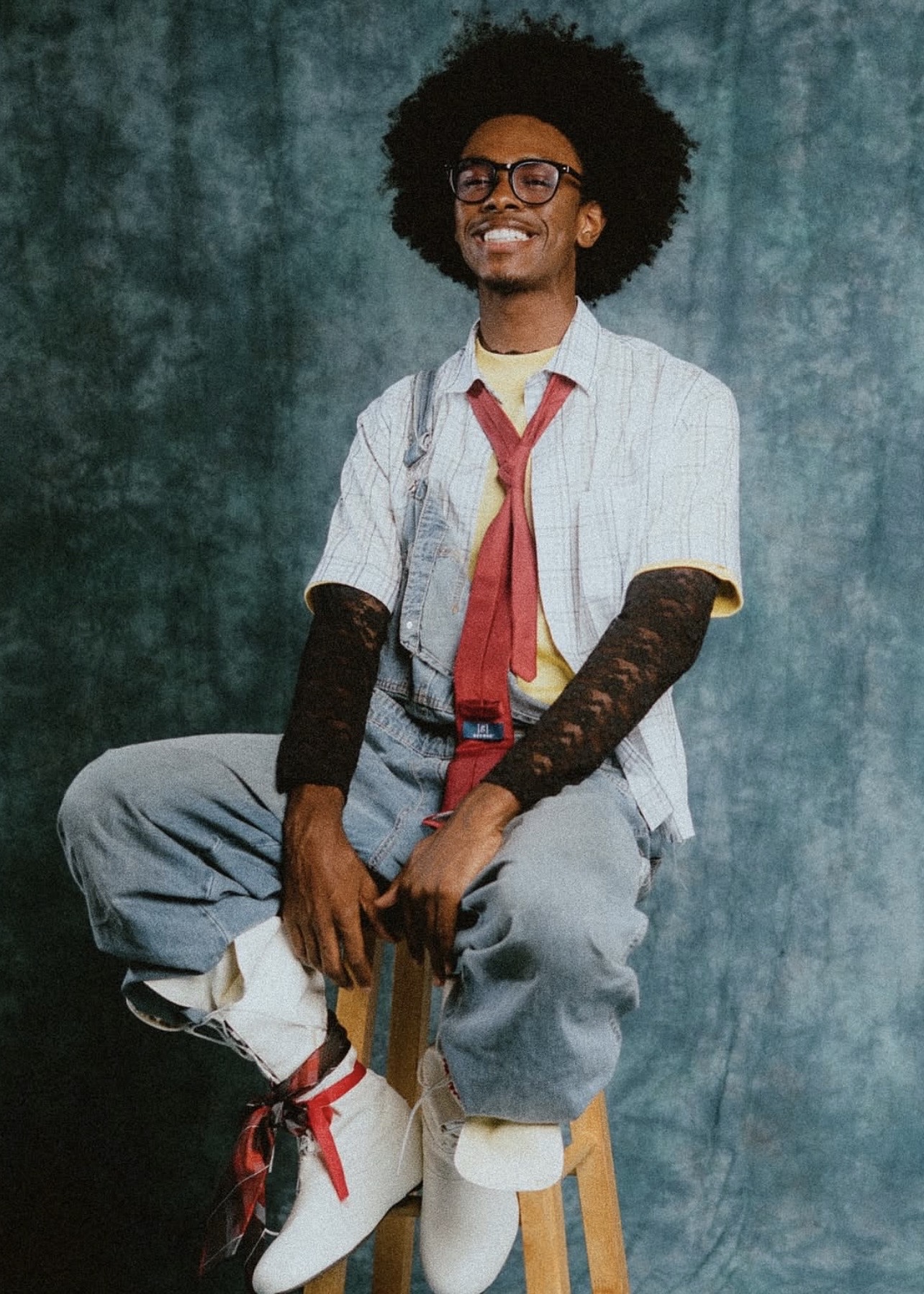 Person with curly hair, glasses, smiling, sitting on a stool, wearing a white shirt, red tie, and gray pants.