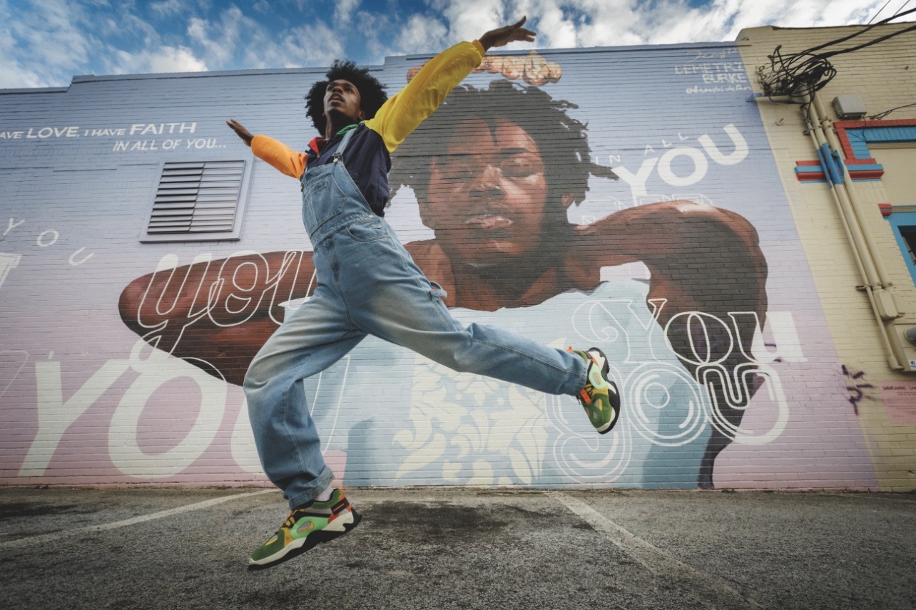 Person jumping in front of a large mural of a woman's face on a building wall, with arms outstretched and legs apart.