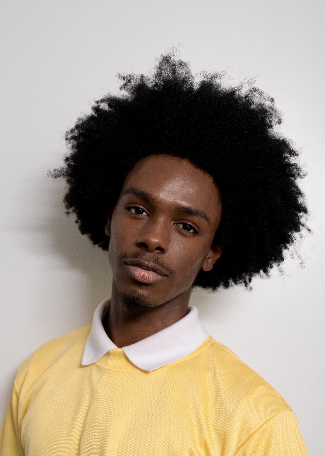 Young man with natural afro hairstyle wearing a yellow shirt and white collar, looking at the camera.