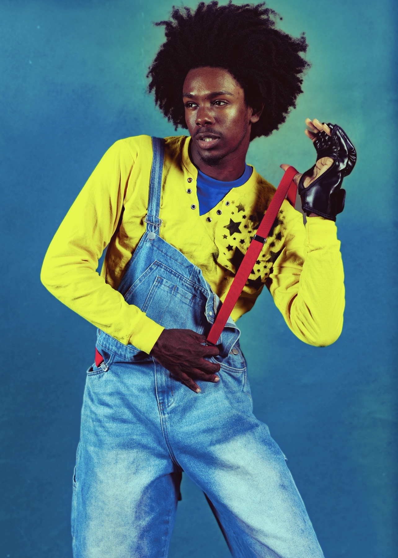 Person with large afro hairstyle wearing yellow shirt and blue overalls holding a red-handled tool against a blue background.