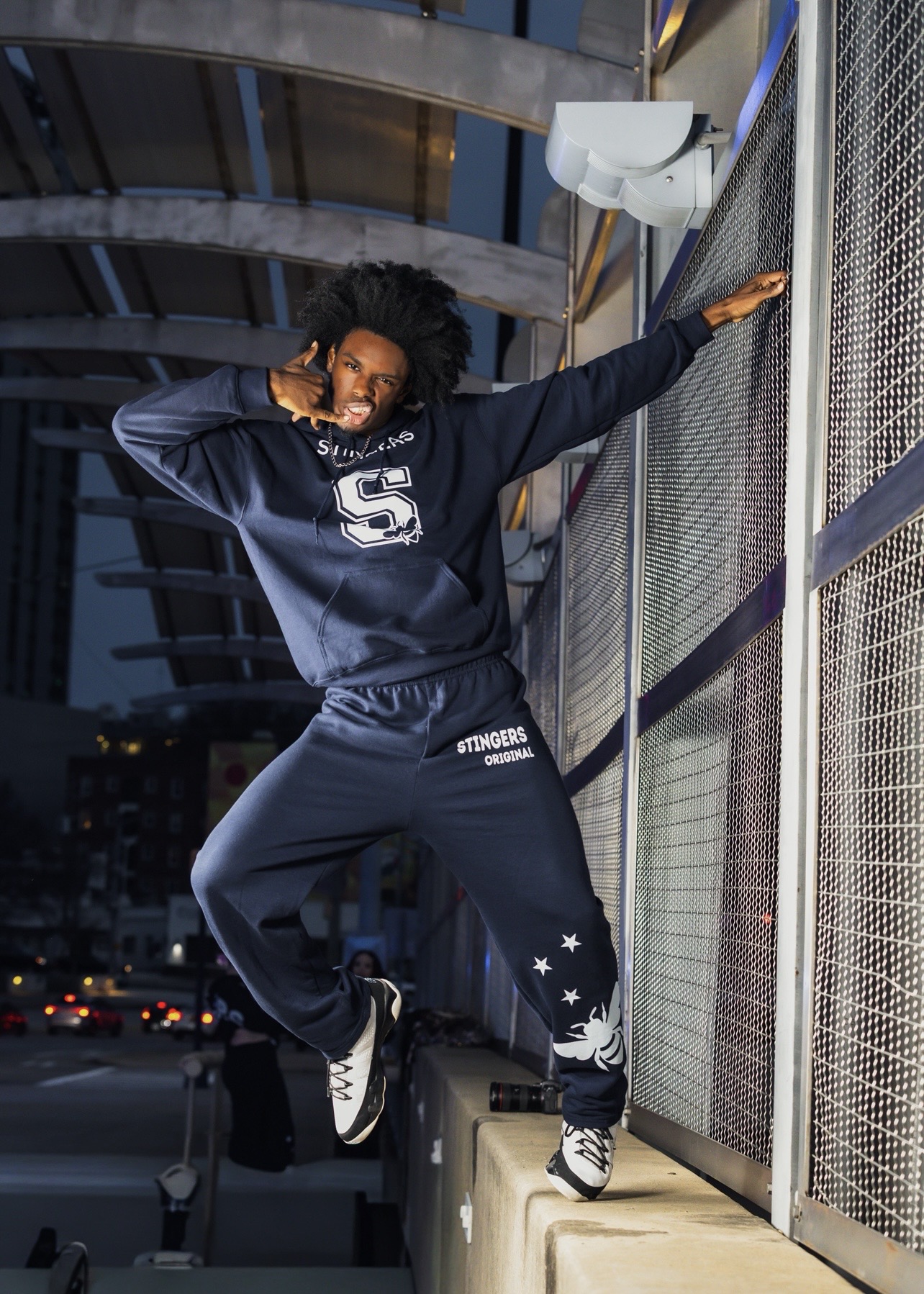 Person with curly hair in a dark tracksuit poses on a ledge outdoors at night, with city lights in background.