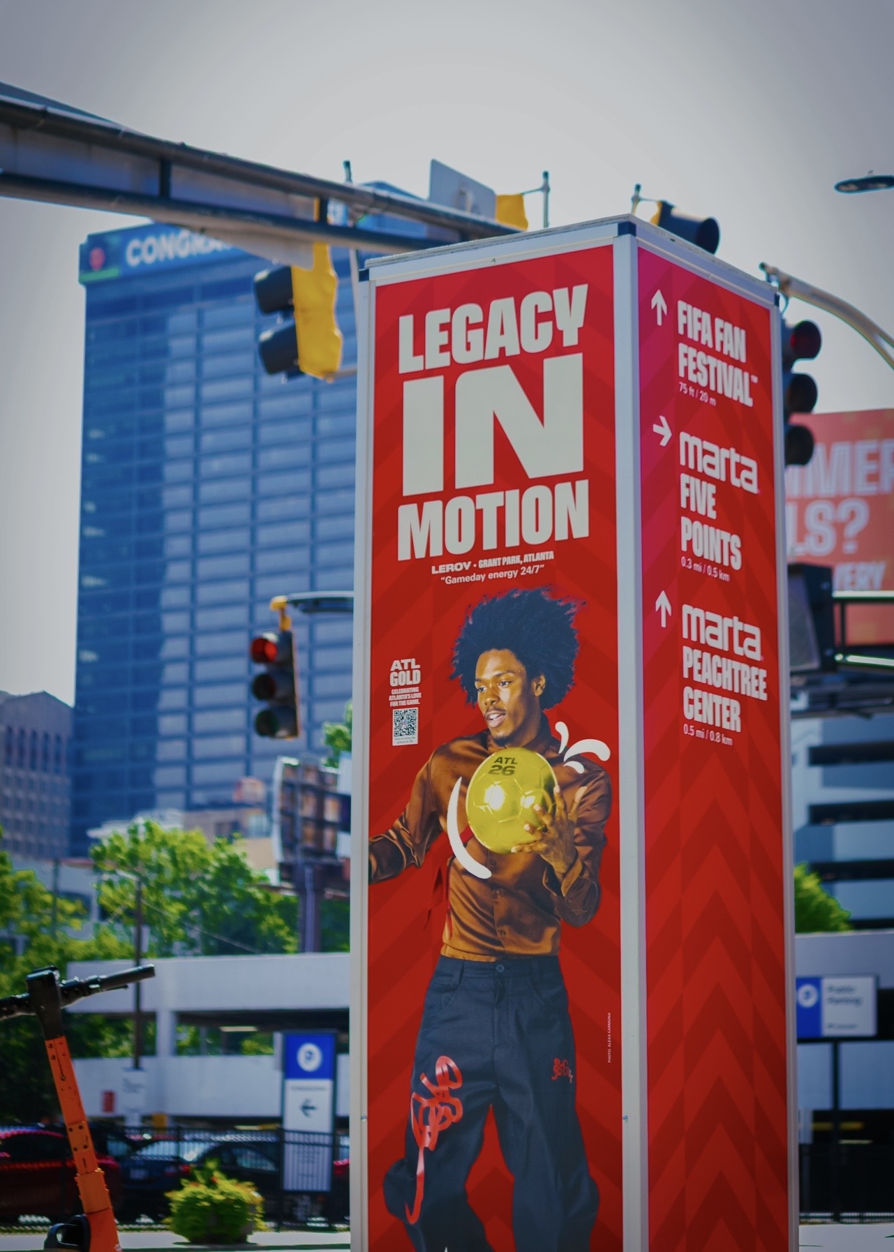 Red vertical sign with white text reading 'Legacy IN Motion' and a person holding a yellow ball, city buildings in background.