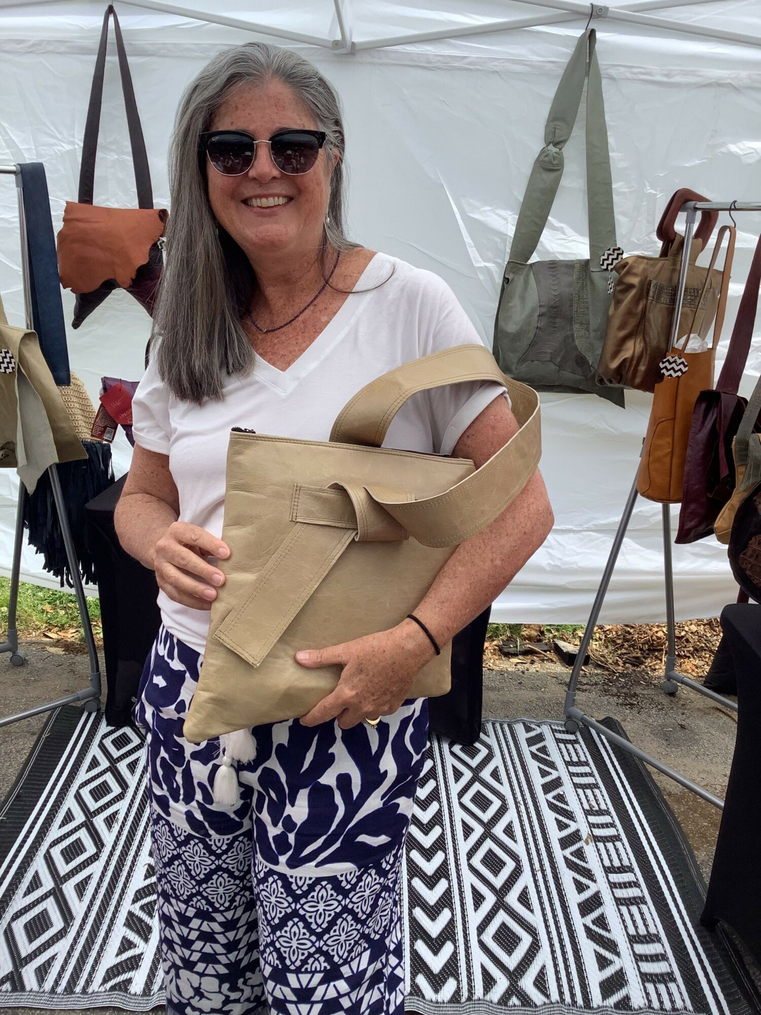 Woman with sunglasses holding a beige shopping bag, standing outdoors near a table with patterned cloth, umbrellas, and clothing racks.