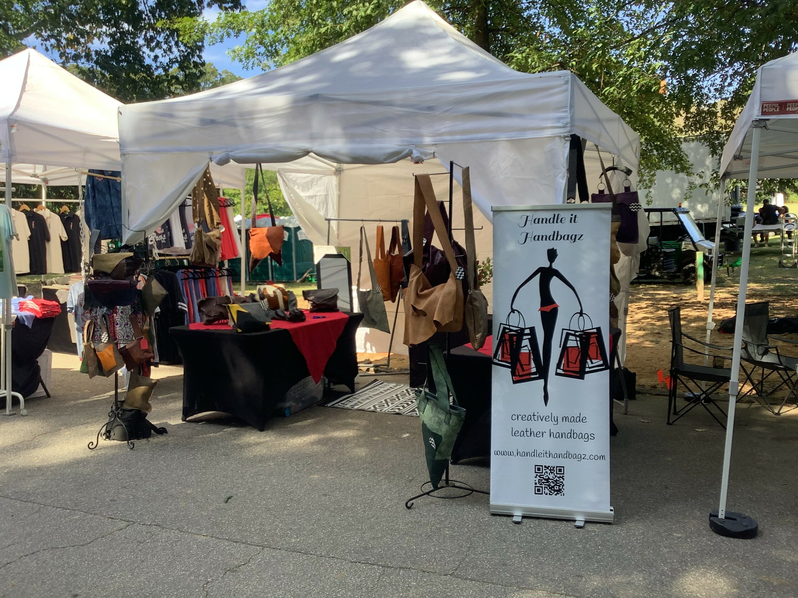 Outdoor market stall with bags and crafts under a white canopy, with a sign displaying a penguin and QR code, trees in background.