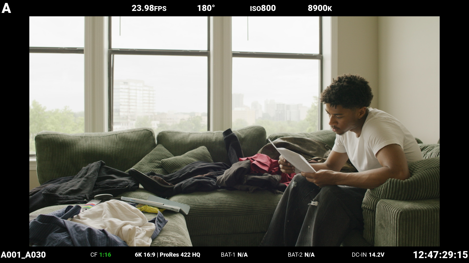 Person sitting on a sofa reading a book in a bright room with large windows, cushions, and scattered items.