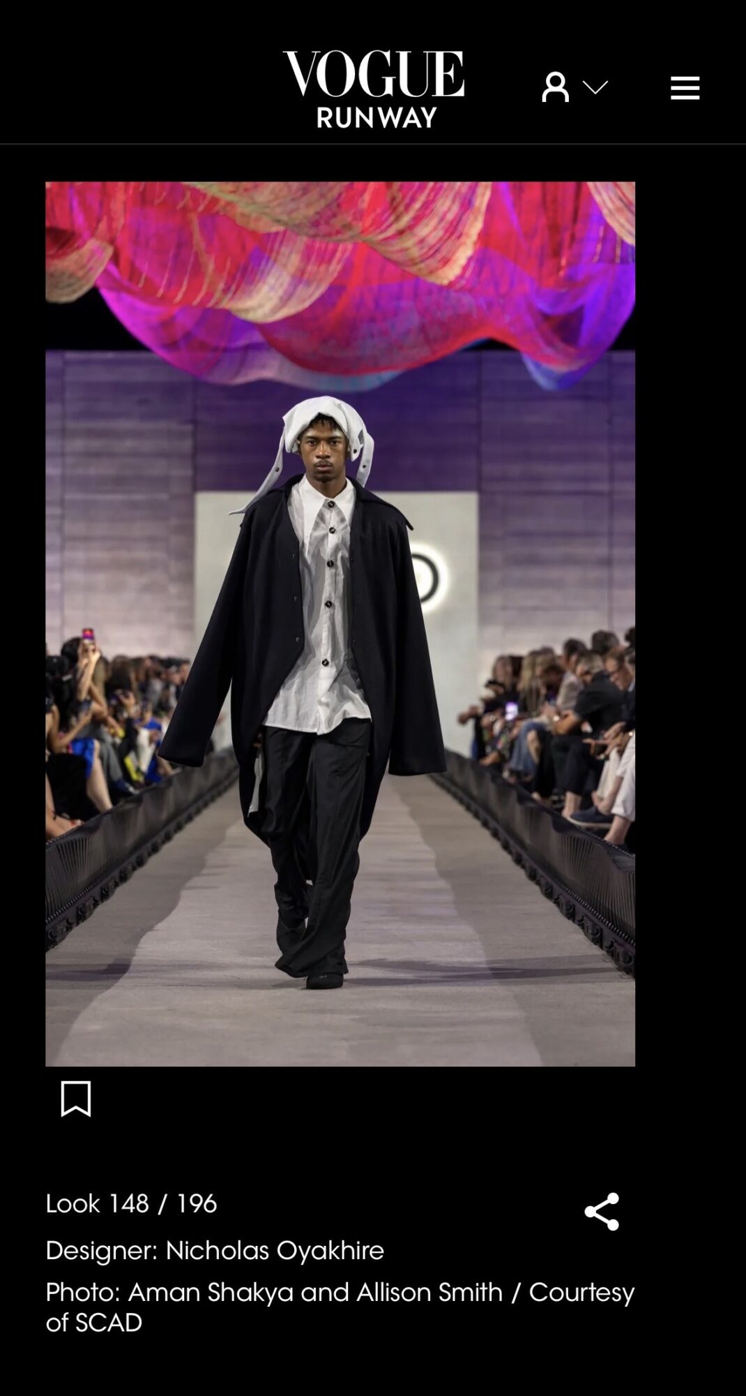 Model walking down runway with audience on both sides, colorful fabric overhead, wearing layered black and white clothing.