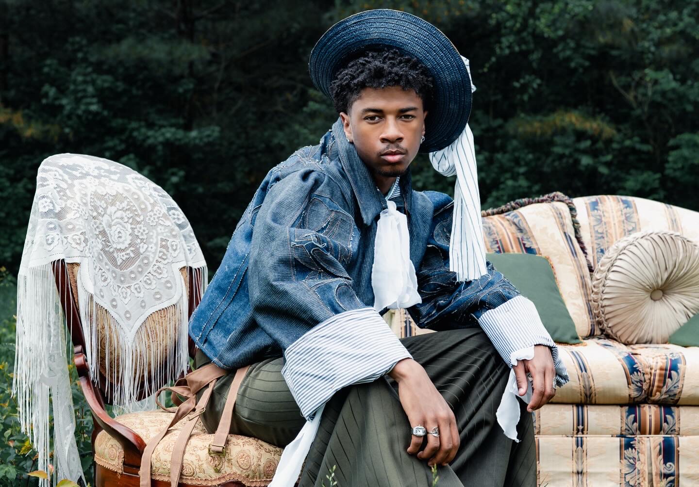 Young man sitting outdoors on a vintage sofa, wearing a large blue hat and denim jacket, with greenery in background.