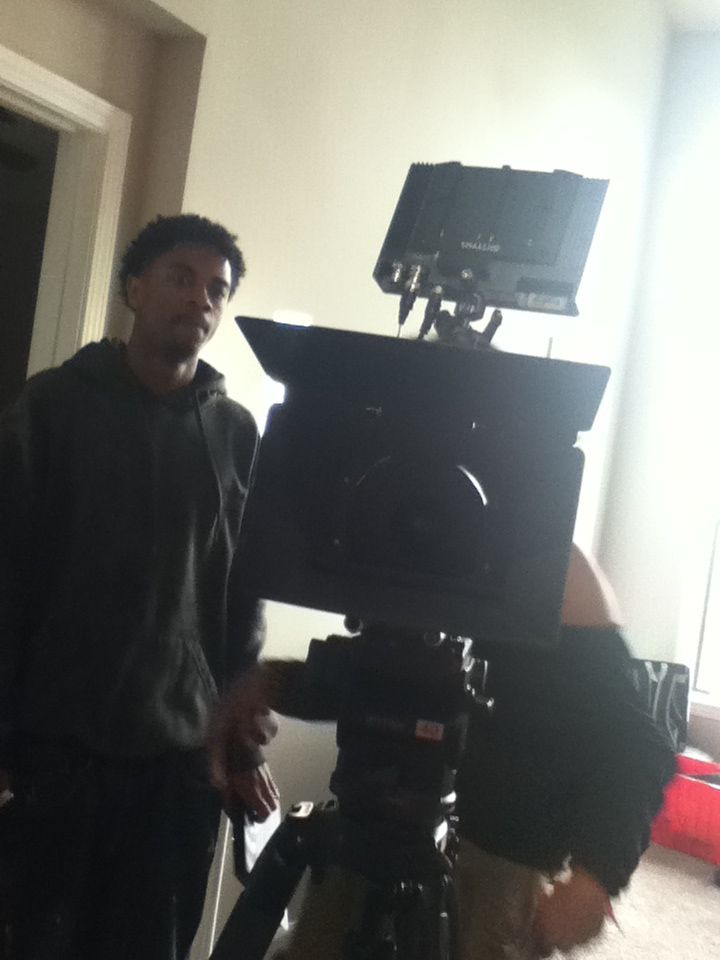 Person standing indoors near camera equipment, with a person holding a phone in the background.