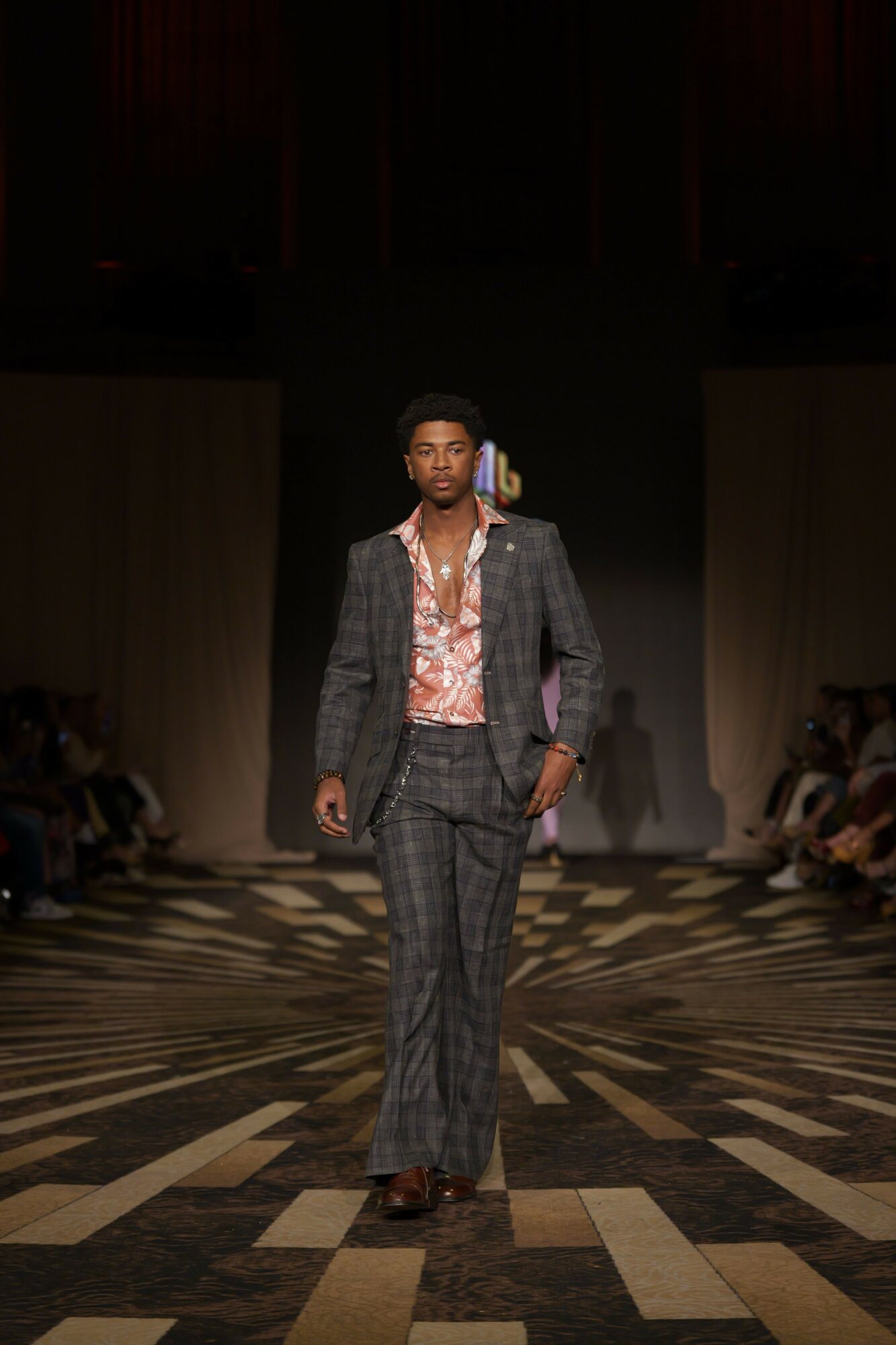 Person standing on a patterned floor, wearing a suit and pink shirt, with a dark background and audience in the distance.