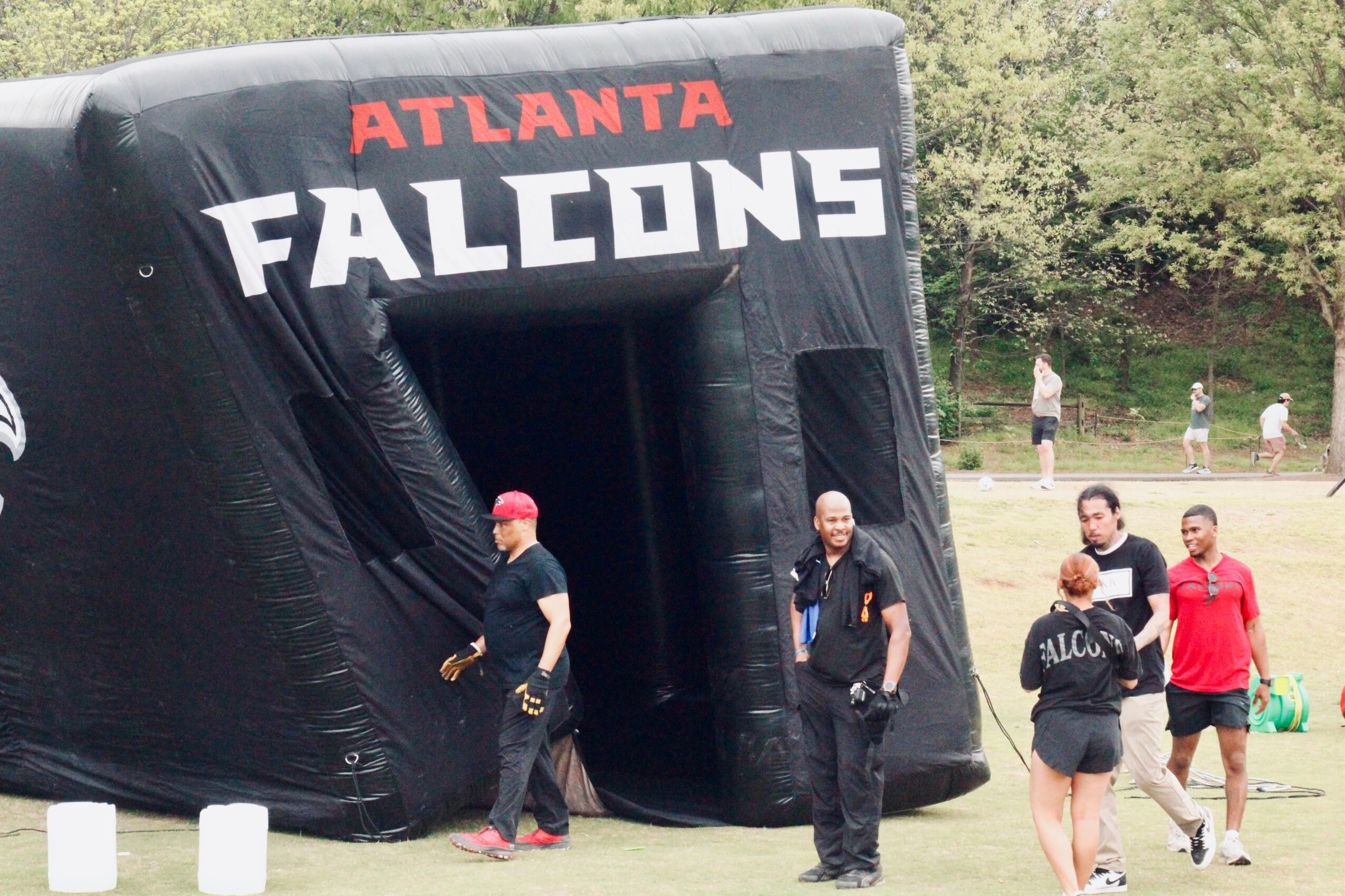 Inflatable tunnel with 'Atlanta Falcons' logo, people walking nearby outdoors, trees in background.
