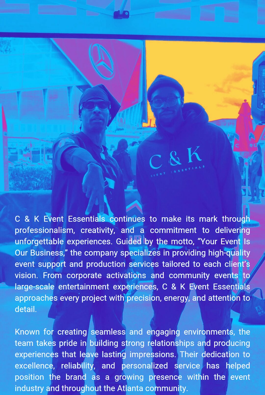 Two people stand outdoors in front of a large screen with a logo, under a colorful sky. One wears sunglasses, the other a cap.