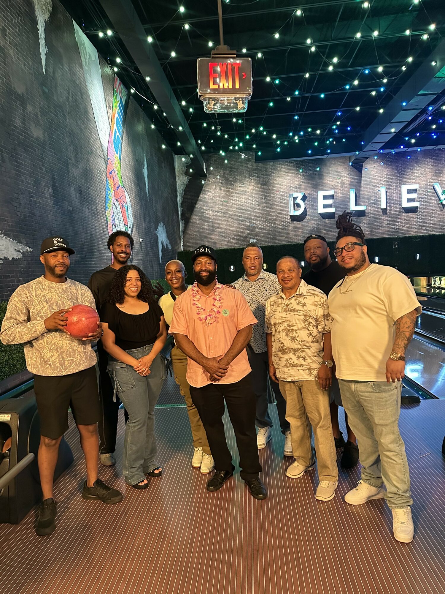 Group of seven people standing indoors, some holding a red ball, with a wall displaying the word 'BELIEVE' and a colorful mural behind them.