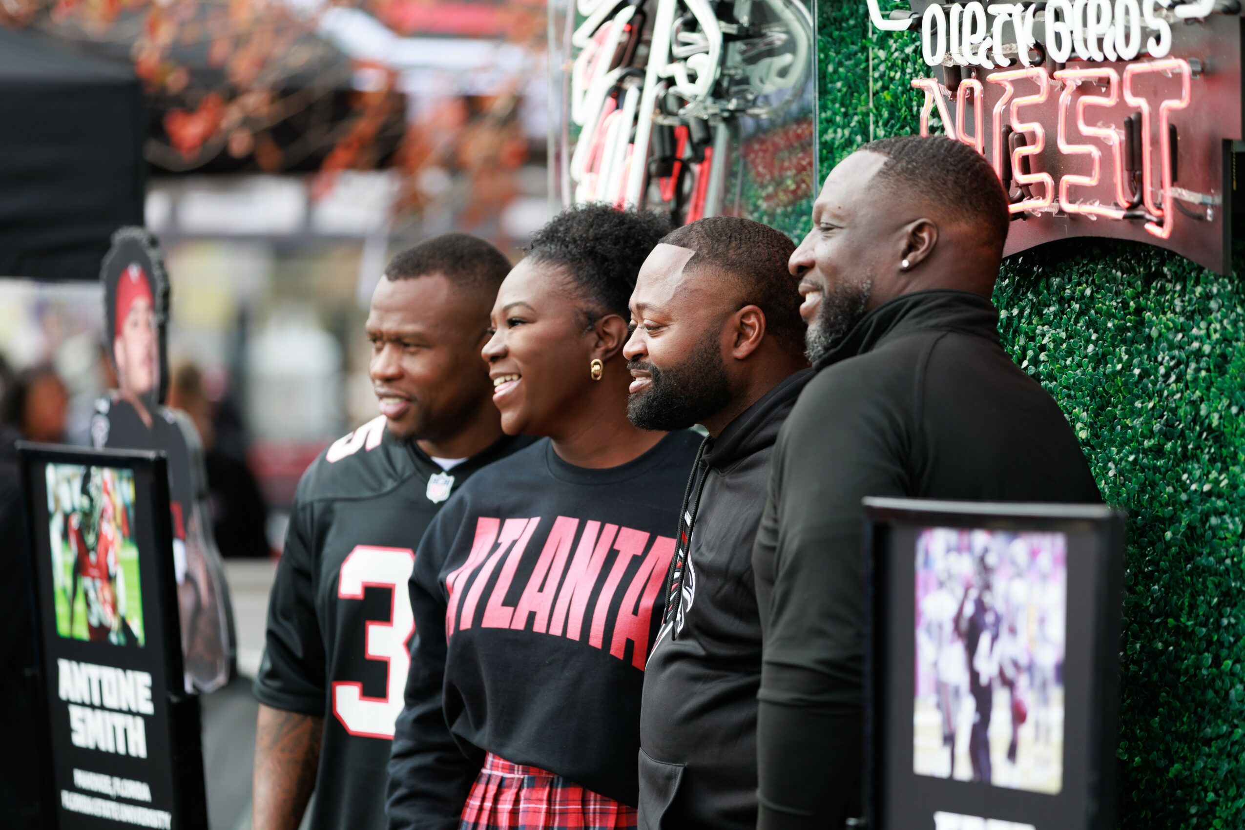 Four people standing together outdoors, smiling, with a green decorated background and a sign that says 'FEST'.