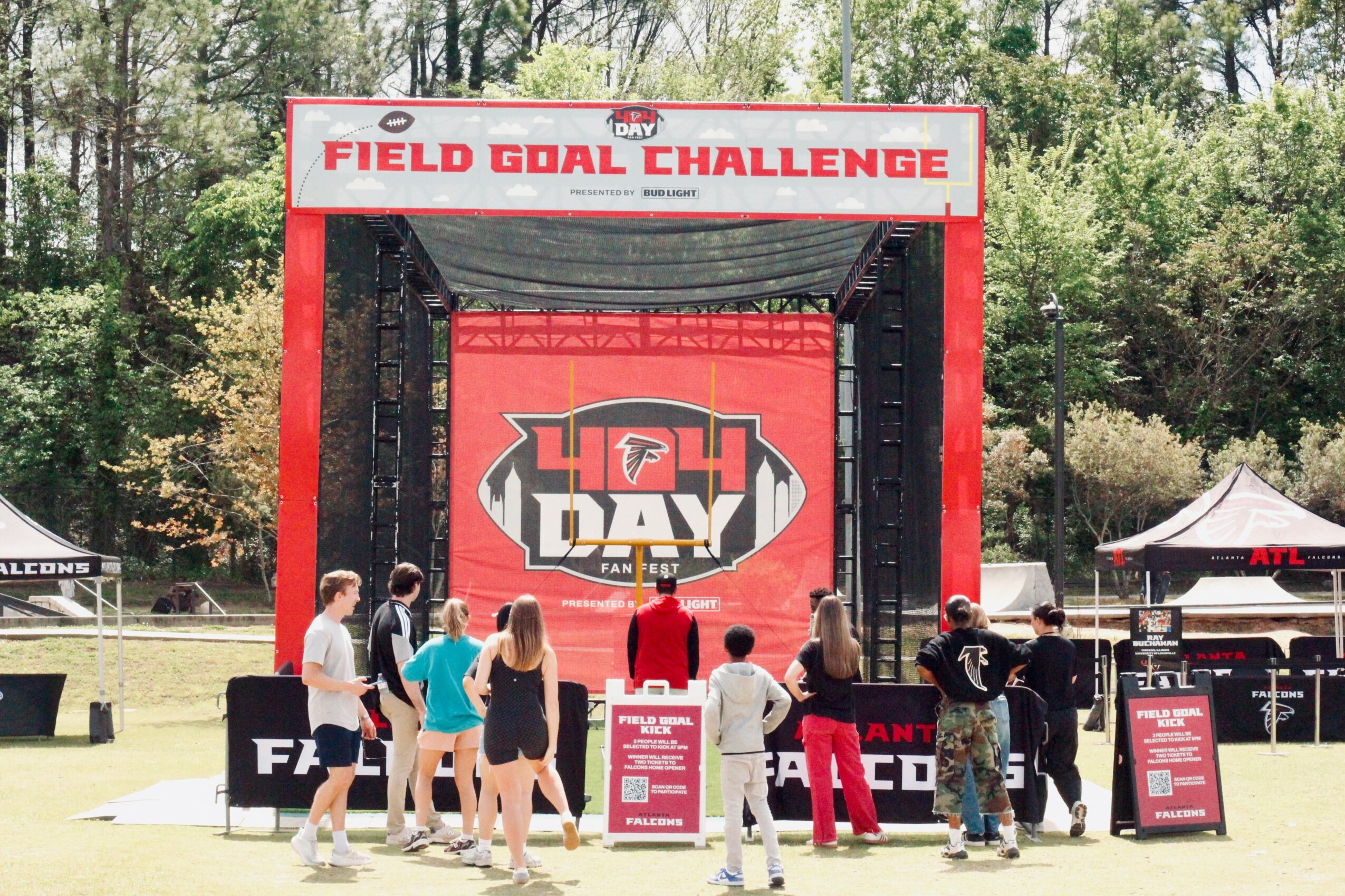 Group of people gathered in front of a stage with a large red banner reading 'Field Goal Challenge' and a logo, outdoors with trees and tents nearby.