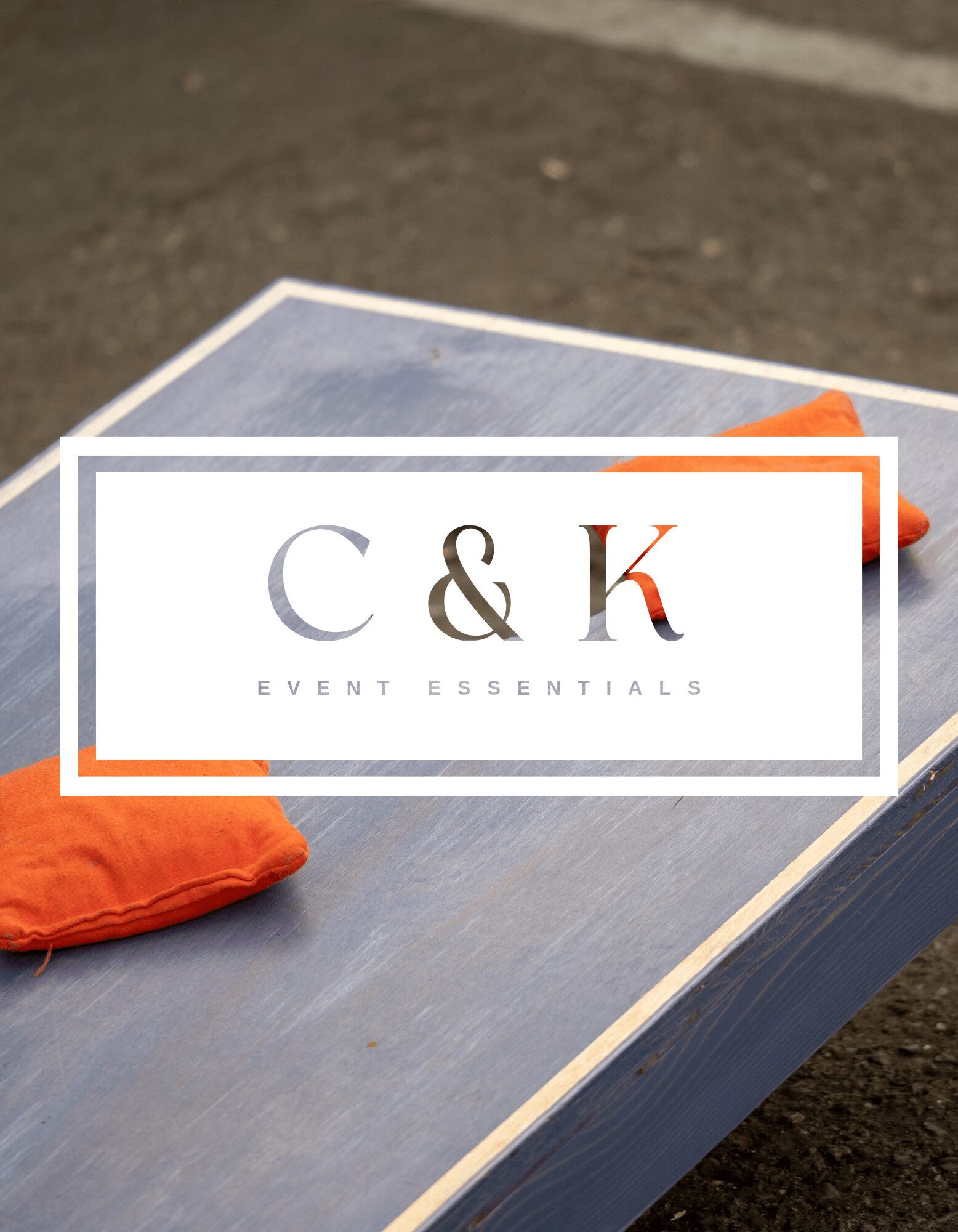 A sign with the letters C and K, and the words EVENT ESSENTIALS, on a table outdoors, with orange cloths nearby.