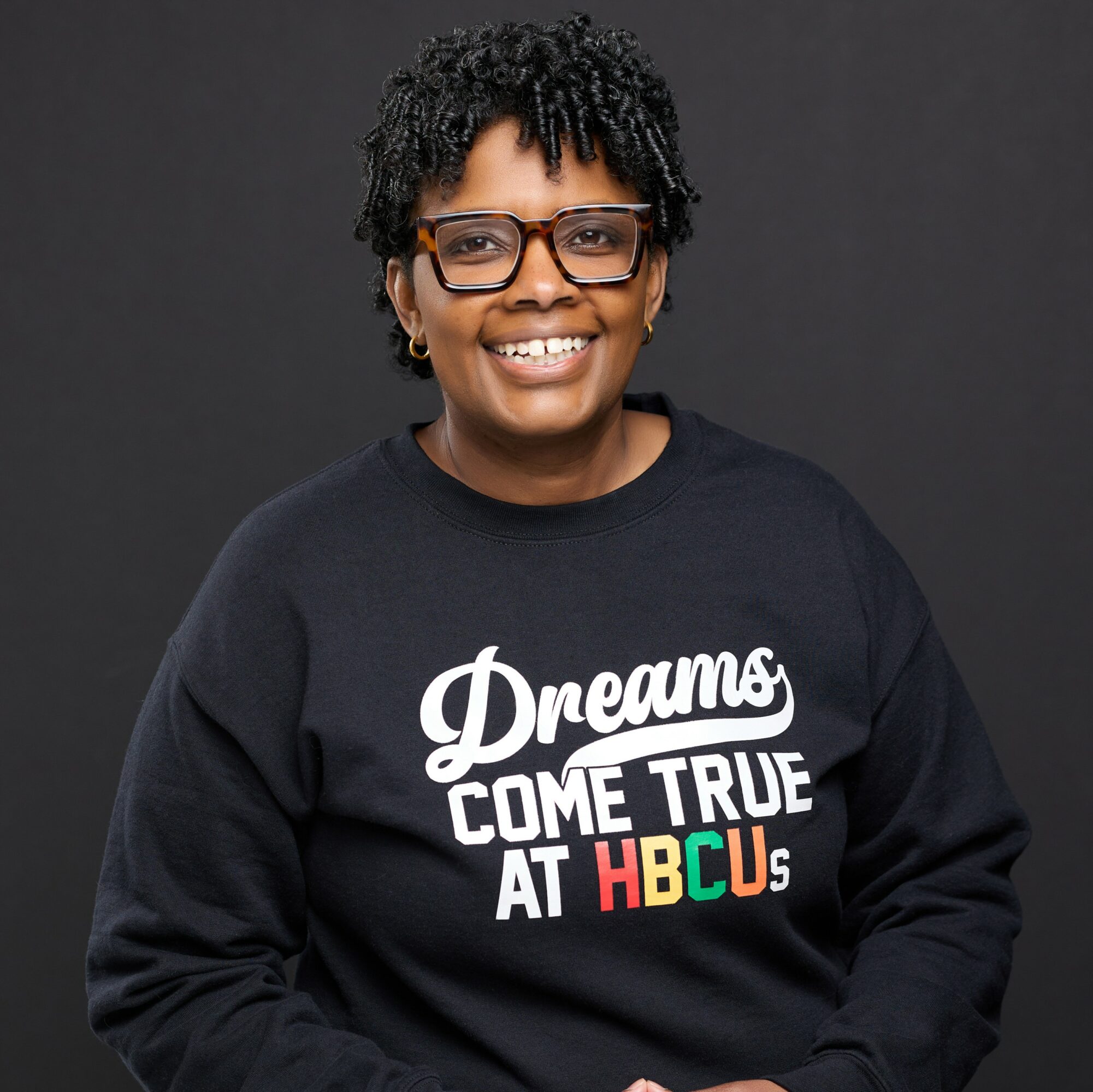 Smiling person with glasses wearing a black sweatshirt with colorful text, standing against a dark background.