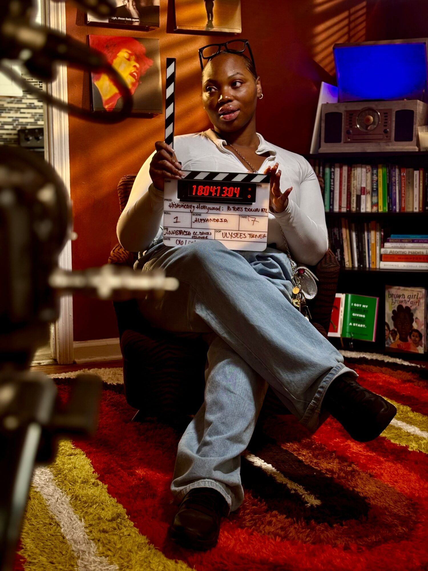 Woman sitting in a room holding a clapperboard, with a bookshelf and colorful rug visible.