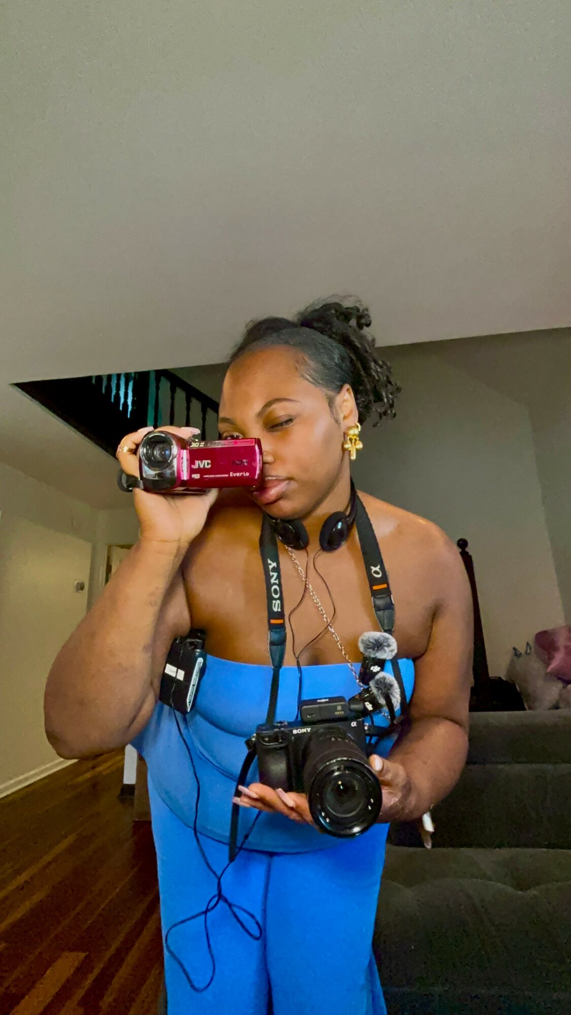 Woman taking a selfie with a camera, wearing a blue dress and headphones, in a room with a gray wall.