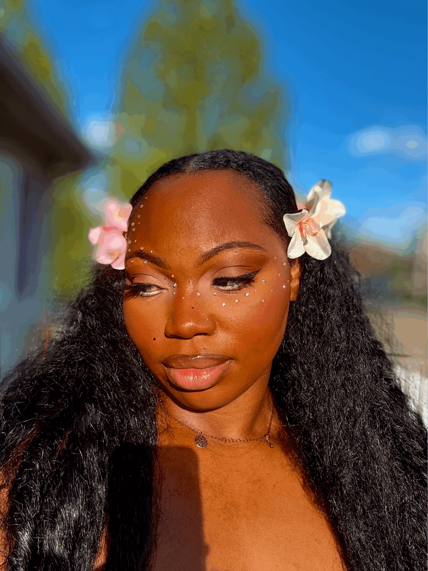 Close-up of a woman with flowers in her hair, outdoors on a sunny day, with a blurred background of trees and sky.