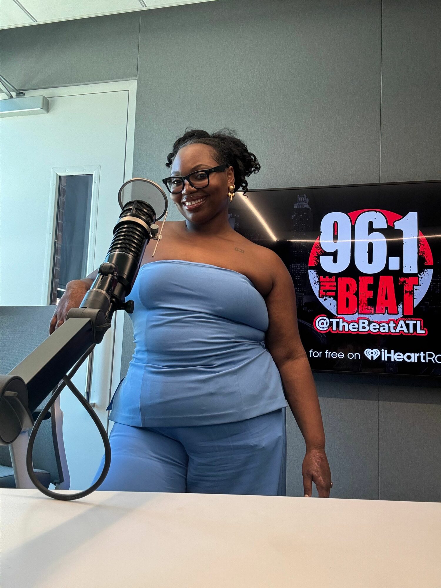 Woman in blue dress speaking into microphone at radio station, with logo in background.
