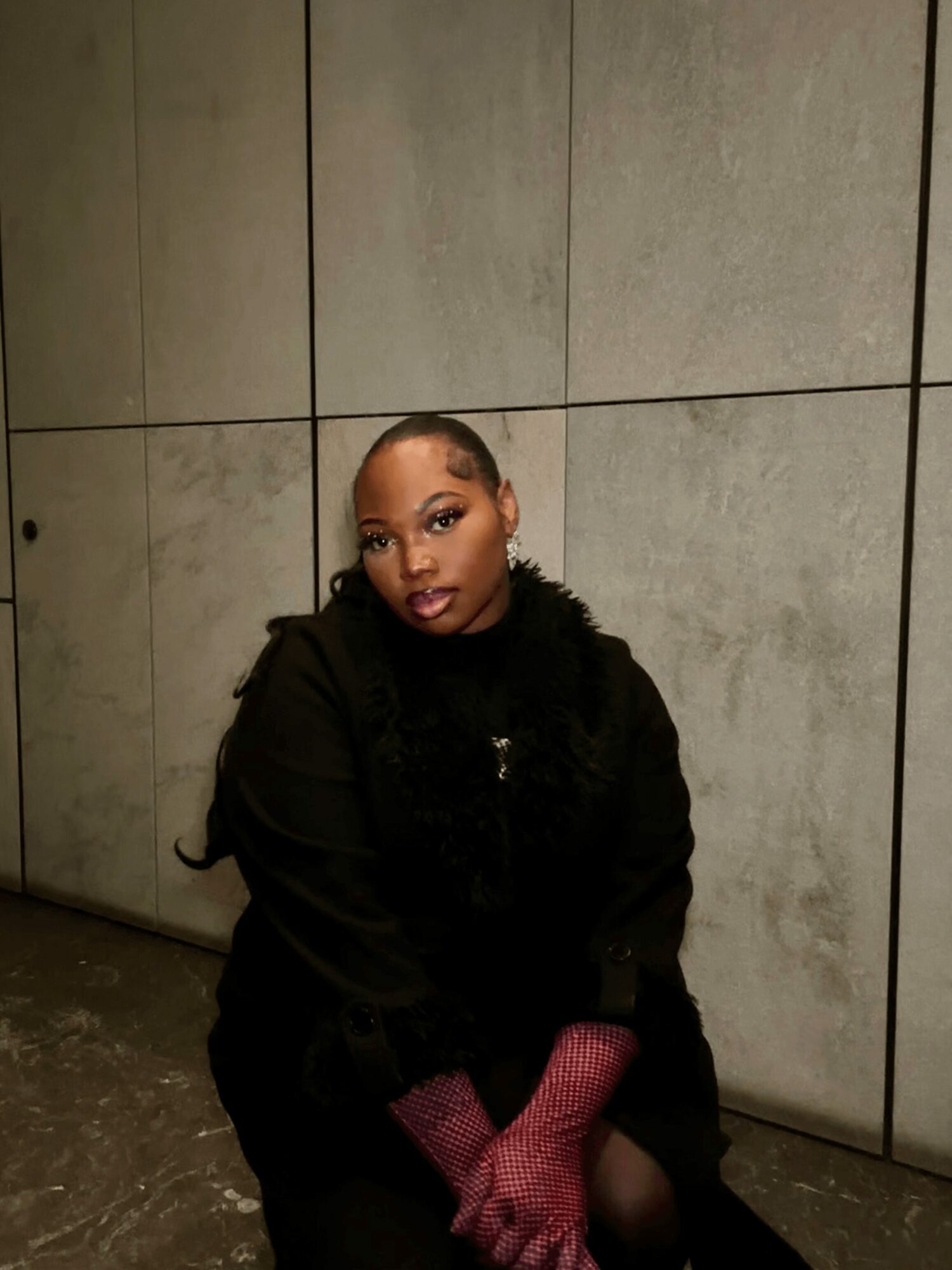 Young woman with short hair and earrings, wearing a black coat and pink gloves, standing against tiled wall.