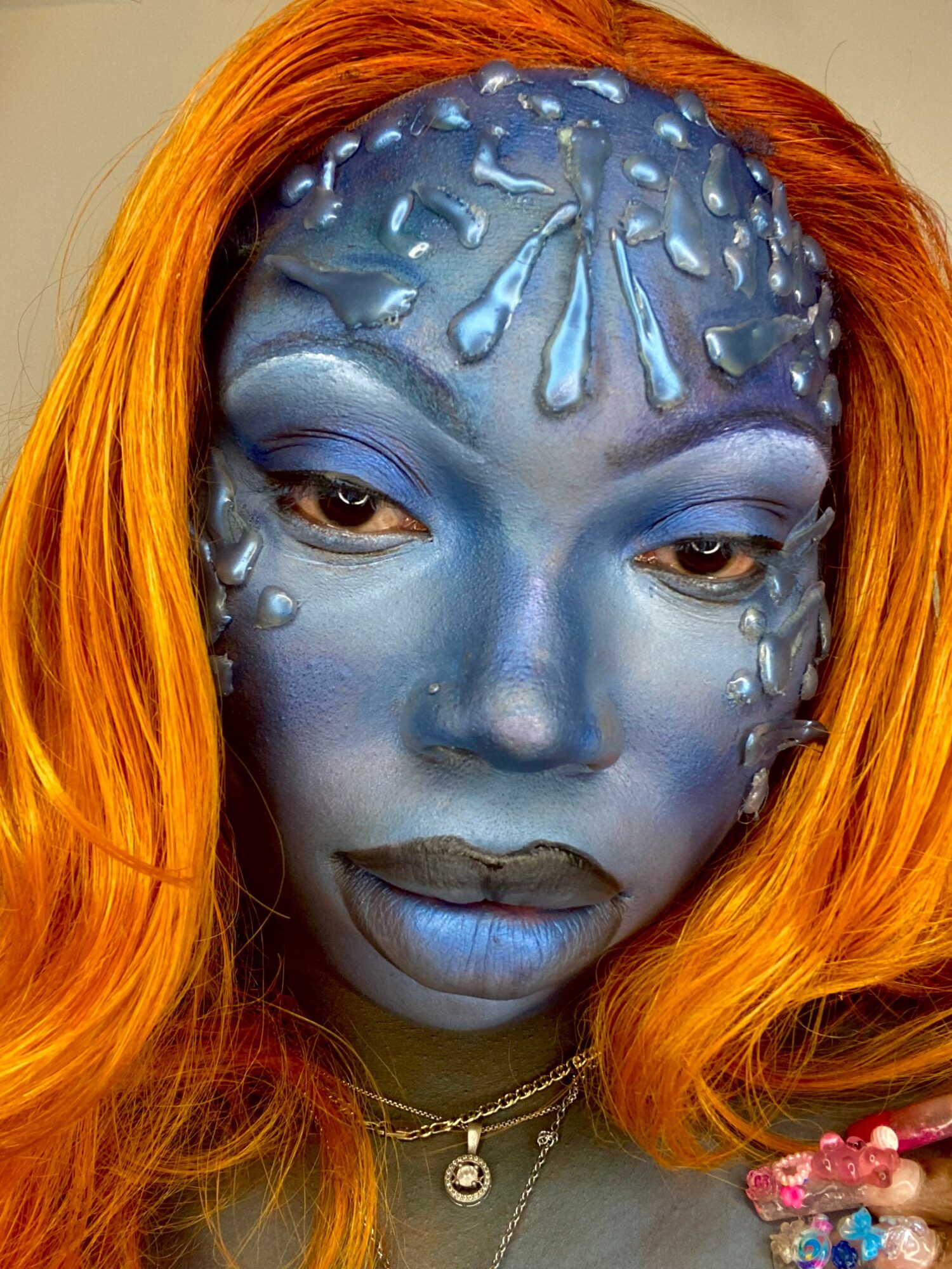 Person with blue face paint, orange hair, and jewelry, looking downward with a neutral expression.
