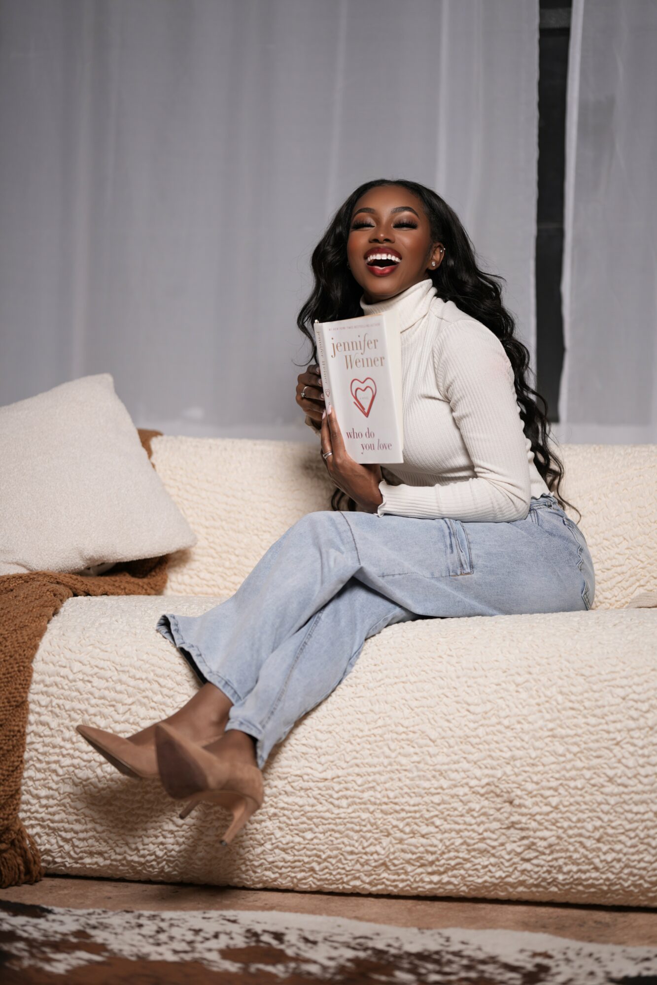 Woman sitting on a sofa holding a notebook with a heart drawing, smiling, wearing a white sweater and jeans.