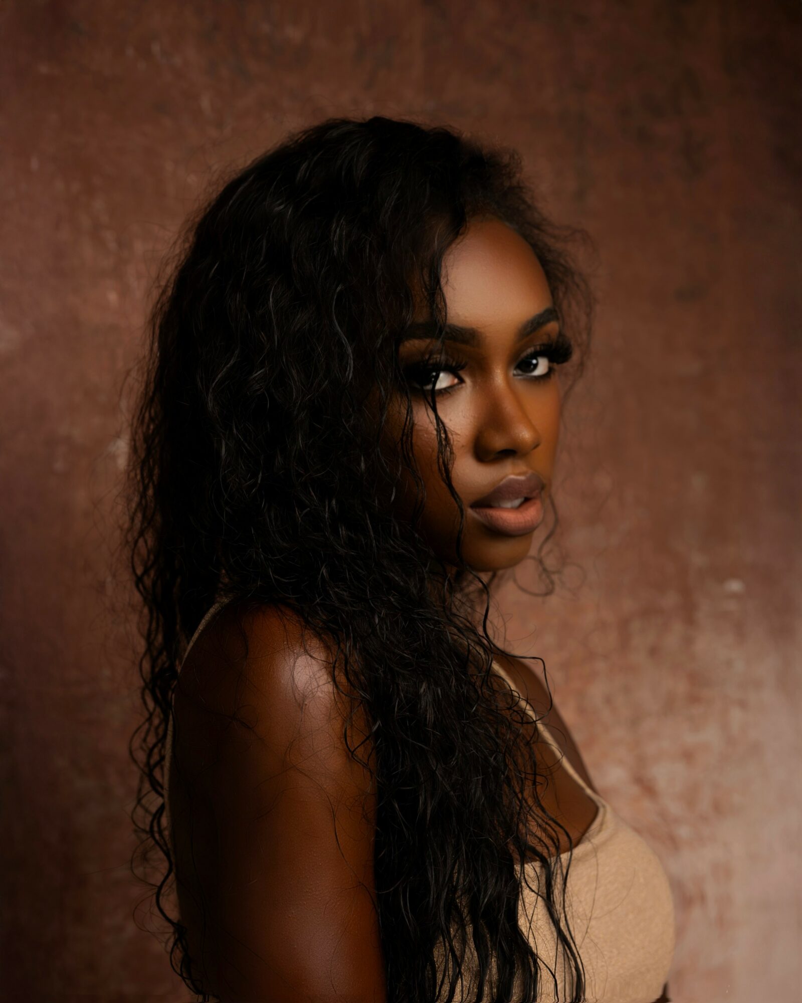 Portrait of a woman with long, curly hair and makeup, looking over her shoulder against a textured background.