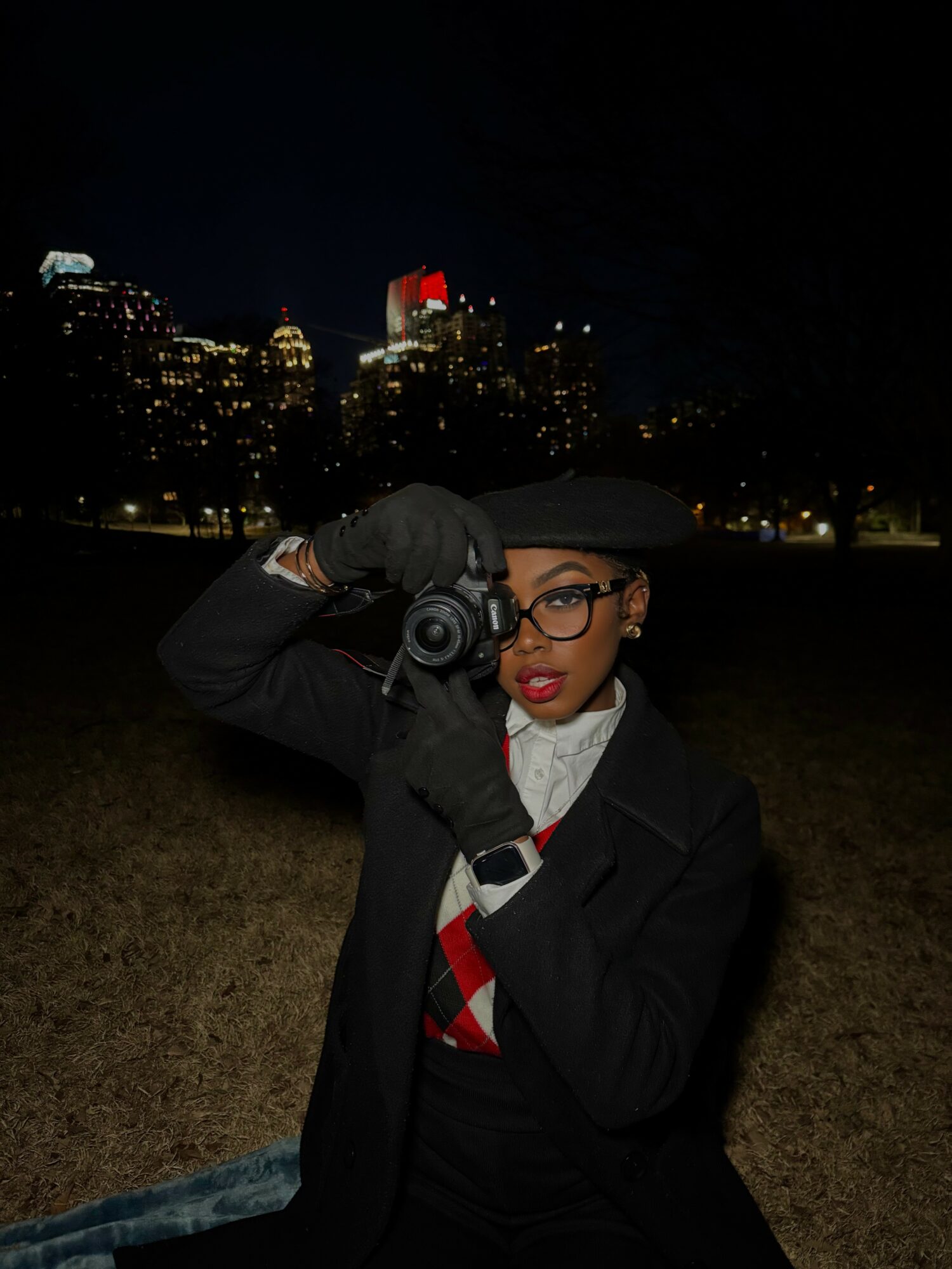 Woman in black coat and hat holding camera, city skyline at night in background, wearing glasses and red lipstick.
