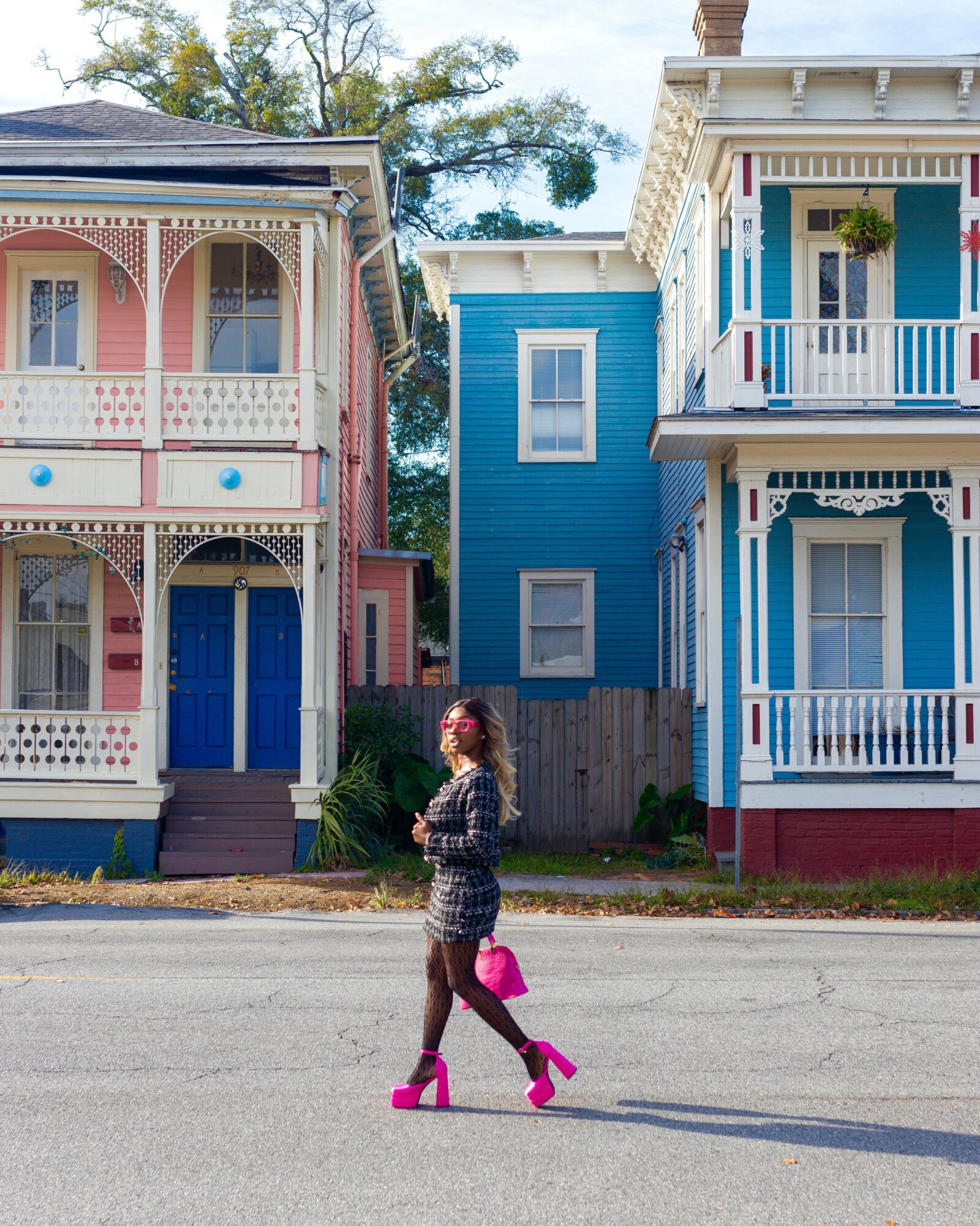 Woman walking on street with colorful houses in background, holding a pink bag, wearing patterned dress and pink shoes.