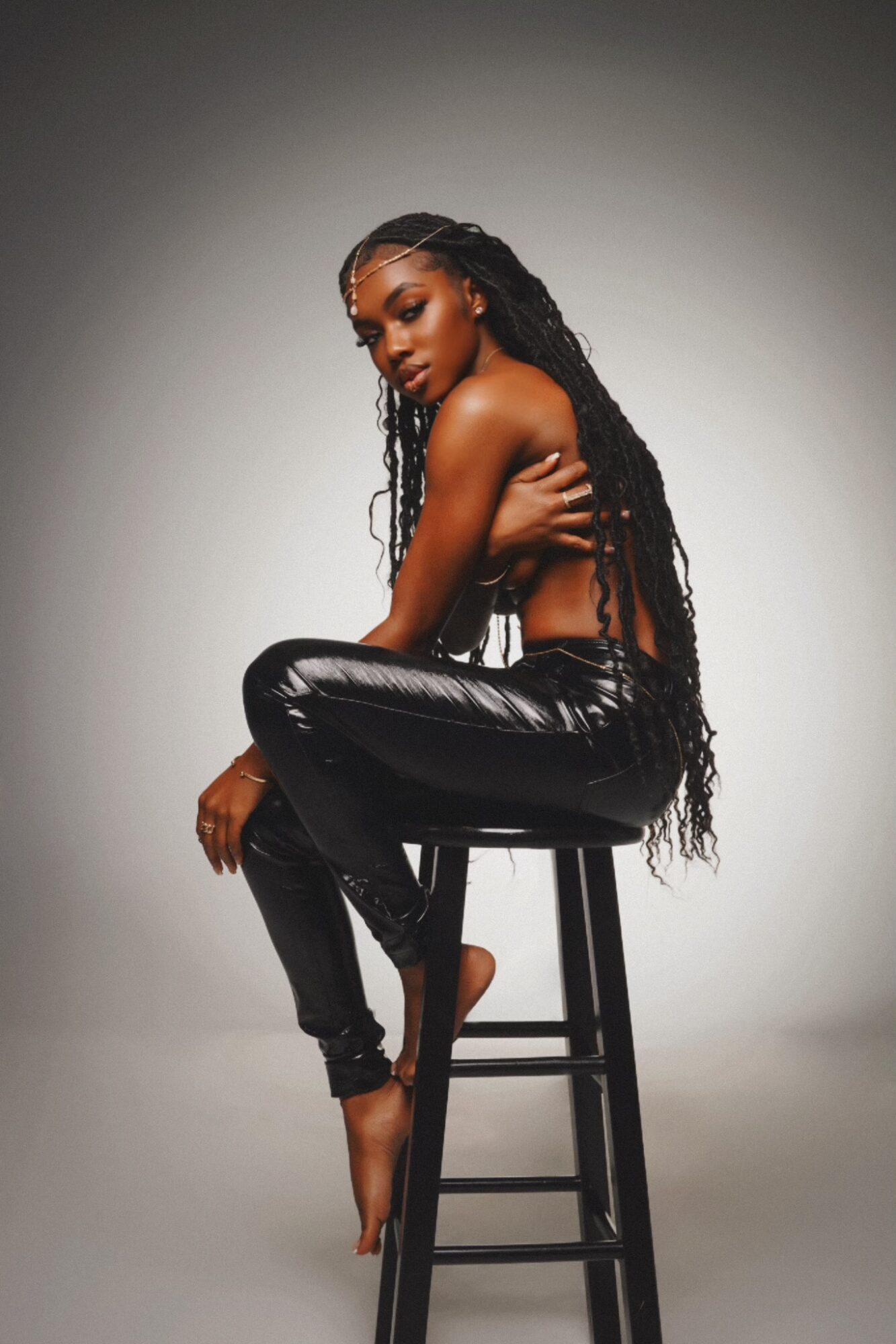 A woman with long, curly hair sits on a stool, wearing shiny black pants and a strapless top, against a plain background.