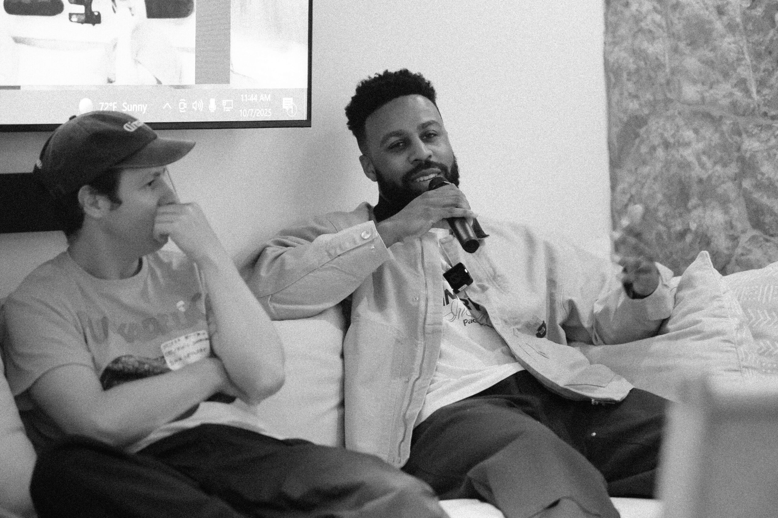 Two people sitting on a couch, one wearing a cap and the other with a beard, talking and smiling.
