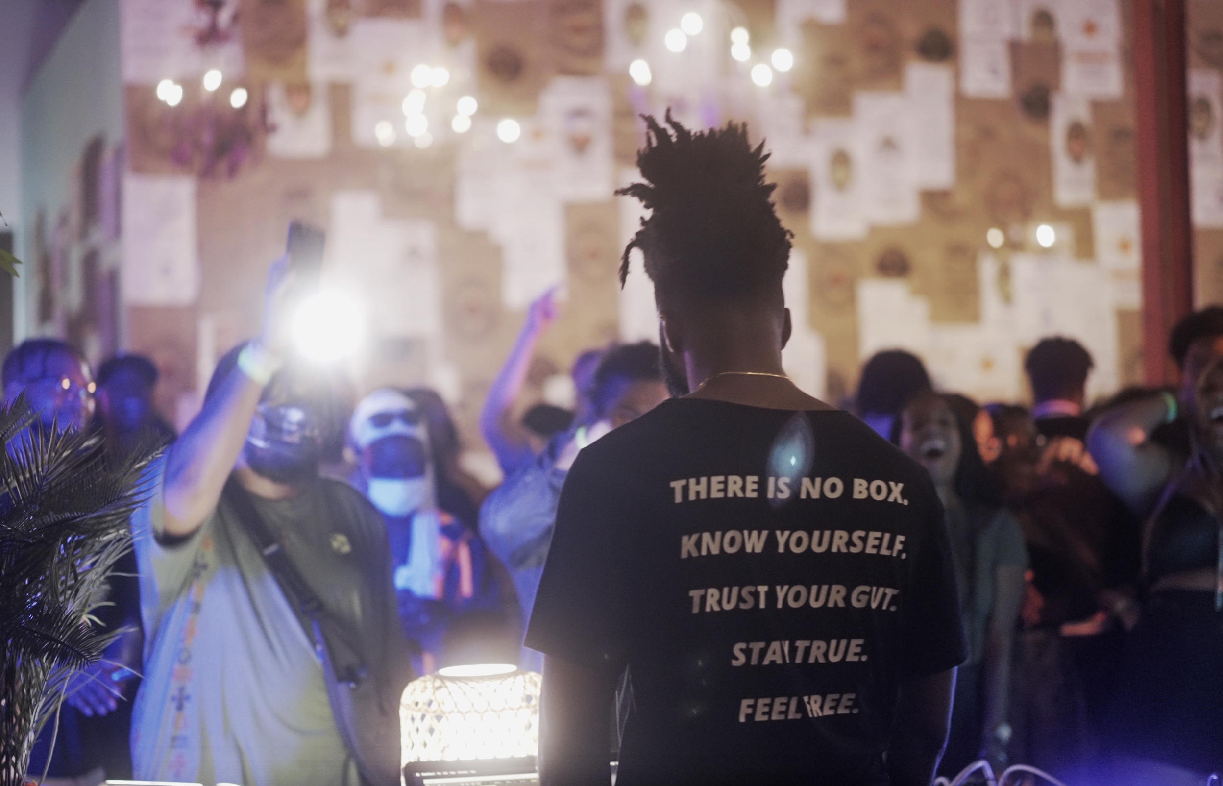 Person with spiky hair wearing a black shirt with white text, surrounded by a crowd in a dimly lit space.