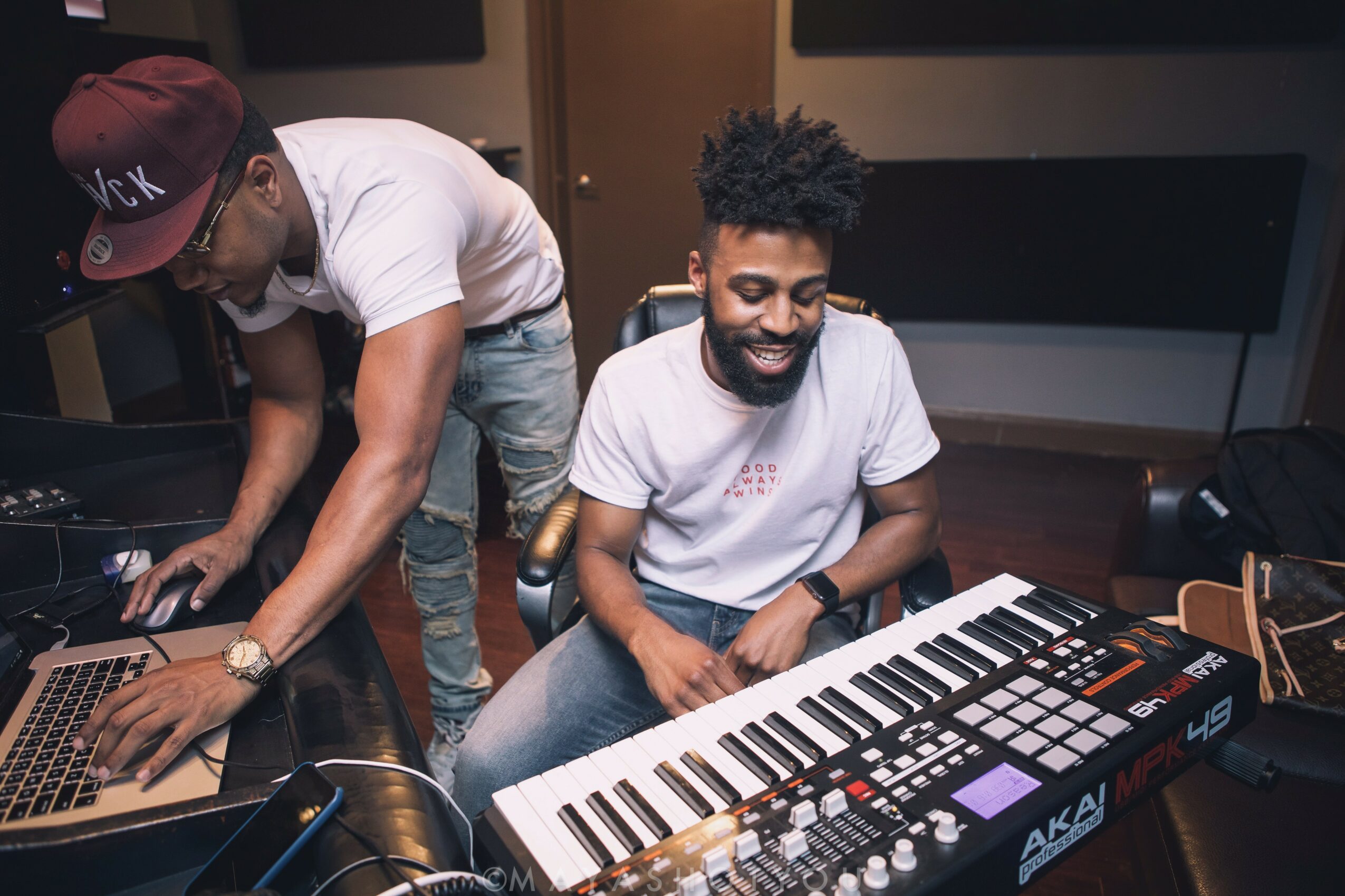 Two men in a music studio, one adjusting equipment and the other sitting with a keyboard and music gear, smiling.