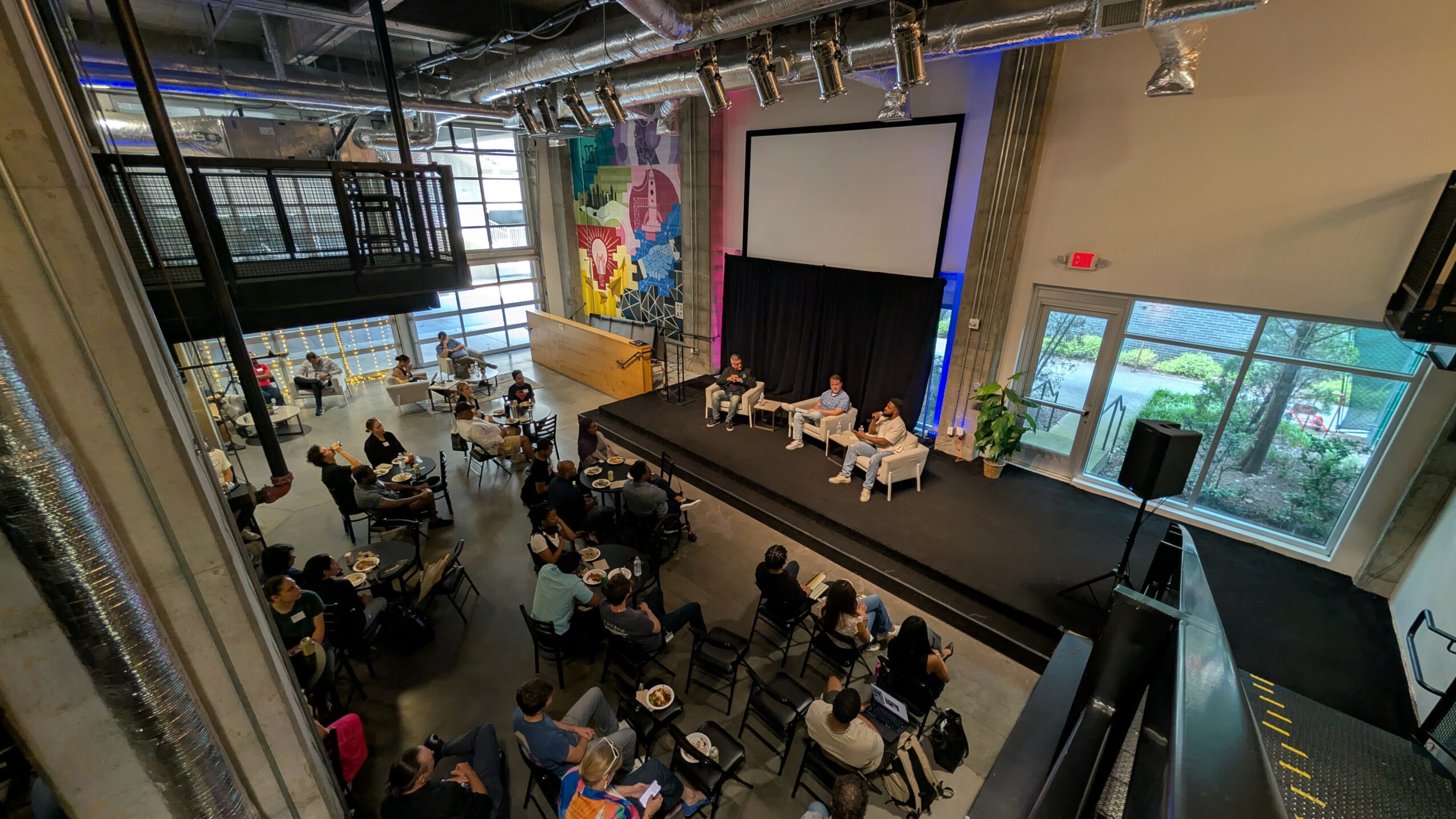 Indoor event space with a stage, panelists, and audience seated at tables, large windows, and colorful artwork on the wall.