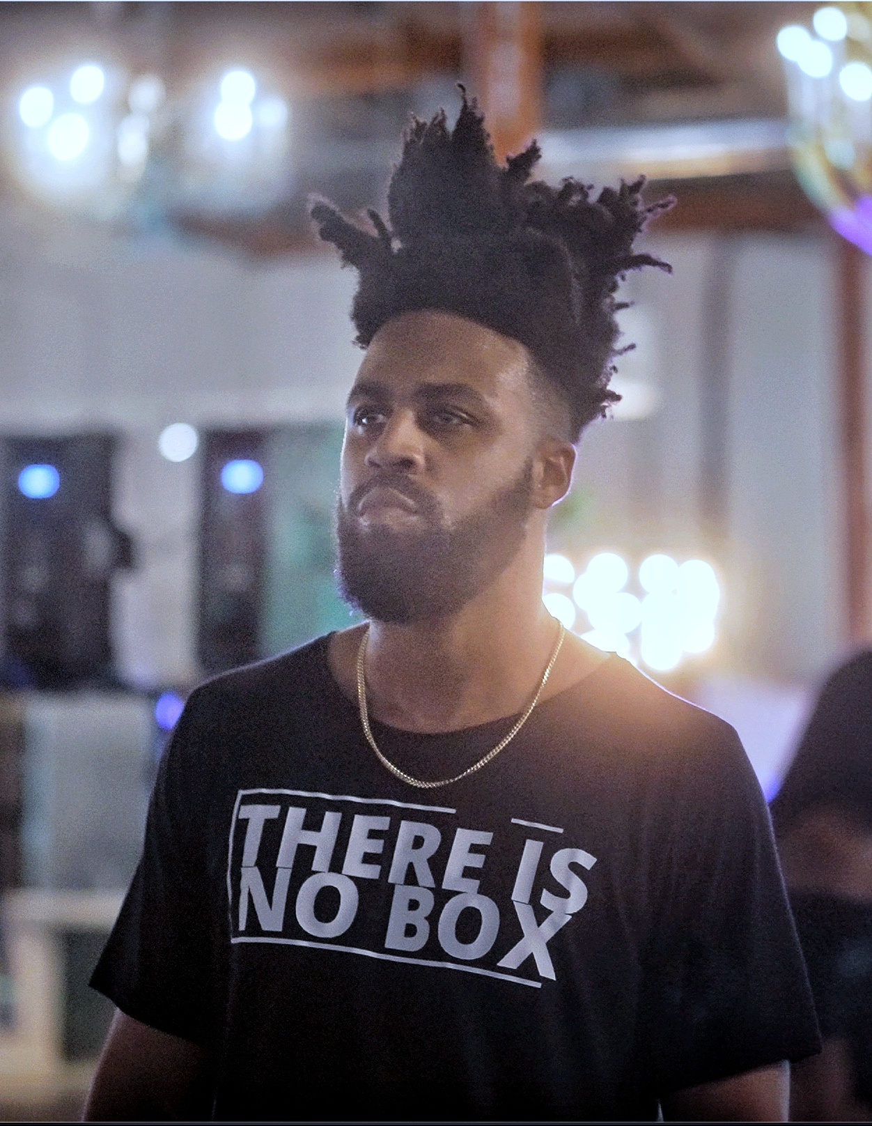 Young man with styled hair and beard wearing a black T-shirt with text, in a dimly lit indoor setting.