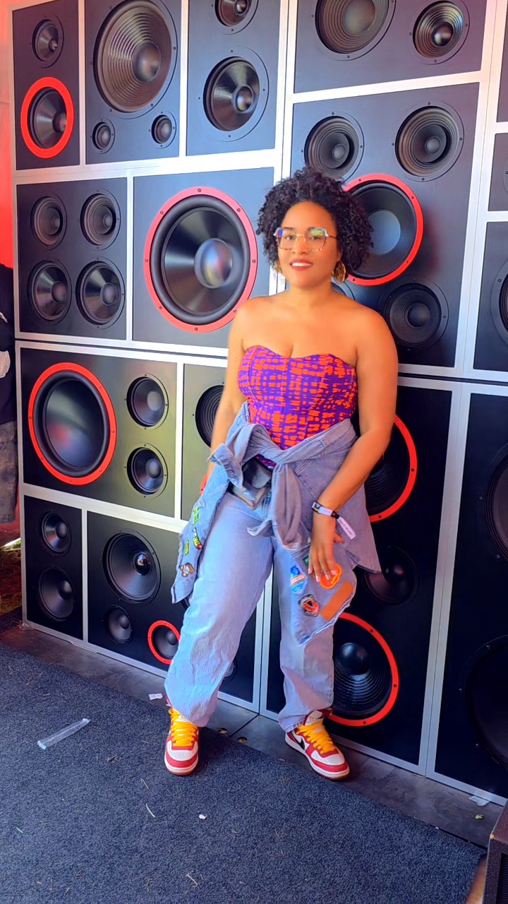 Woman with curly hair and glasses standing in front of a wall of large speakers, wearing a colorful top, jeans, and sneakers.