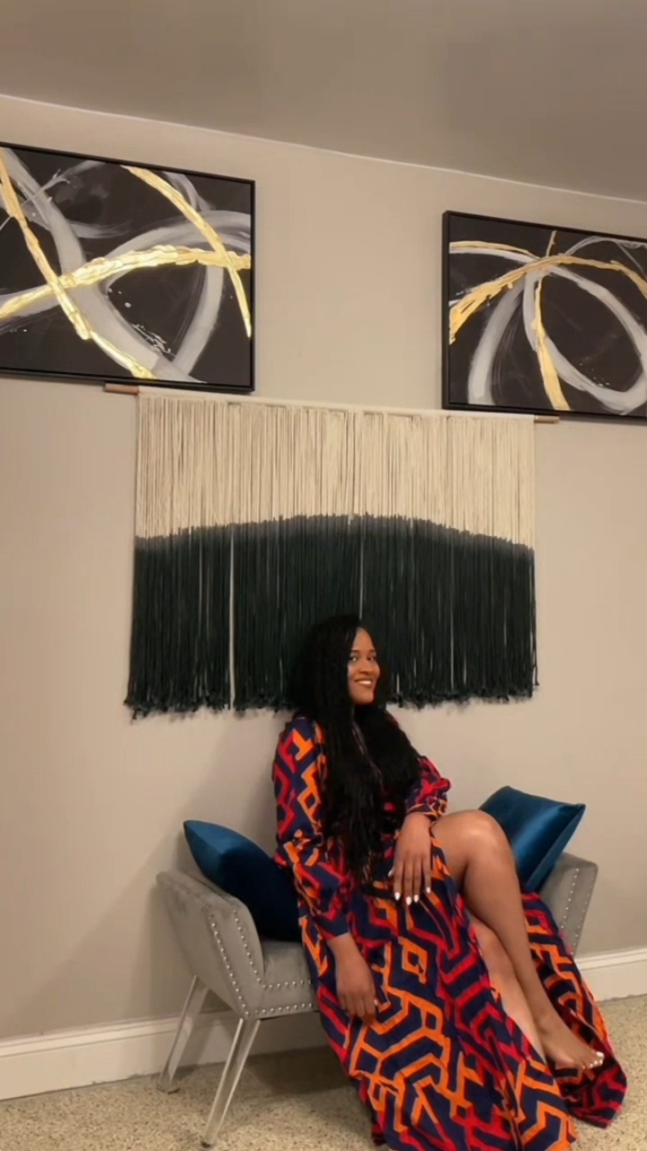 Woman sitting on a chair with a colorful dress, two abstract paintings above, and a decorative wall hanging behind her.