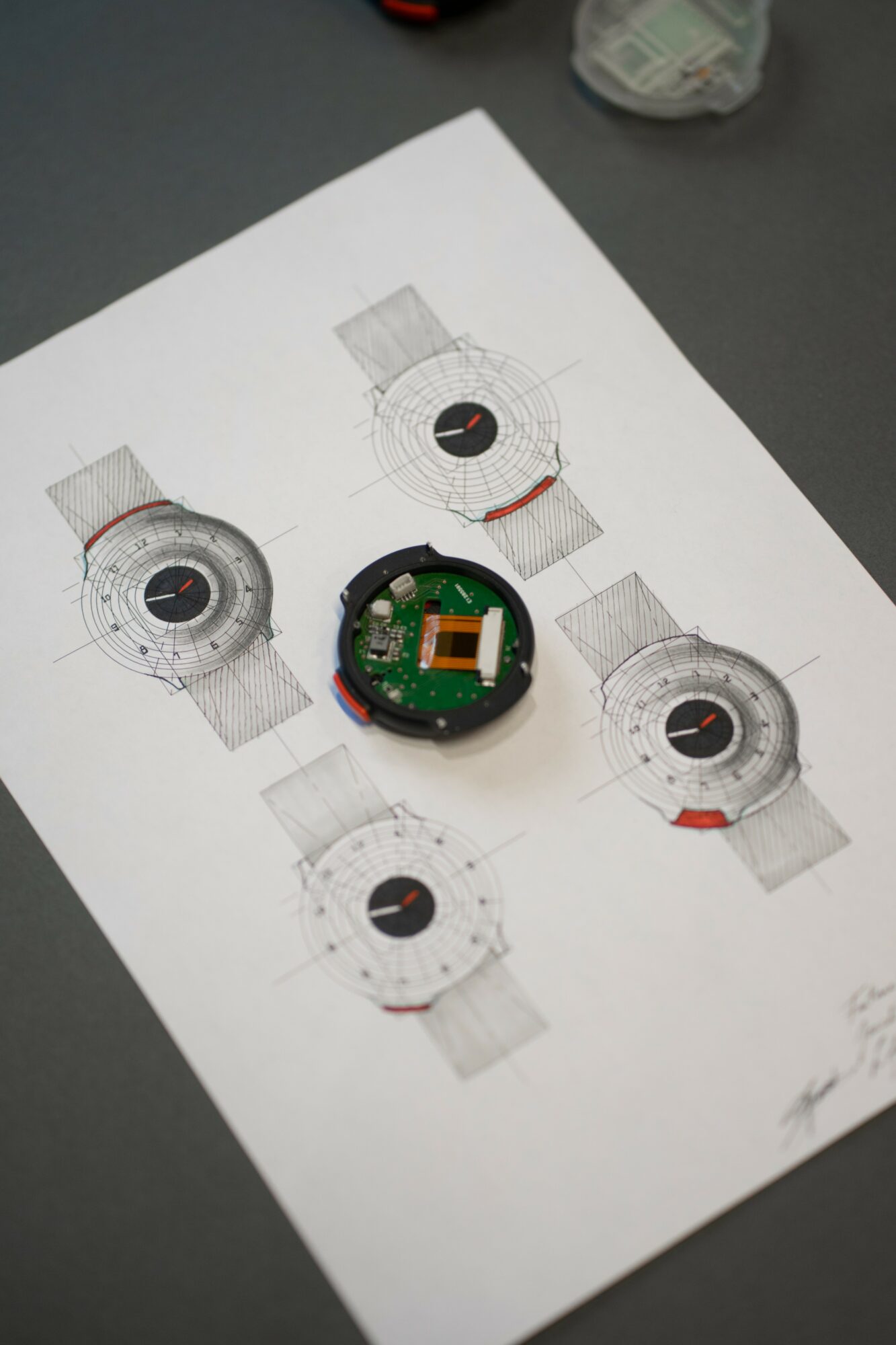 A watch movement placed on a sheet with watch face sketches, on a dark surface with objects in the background.