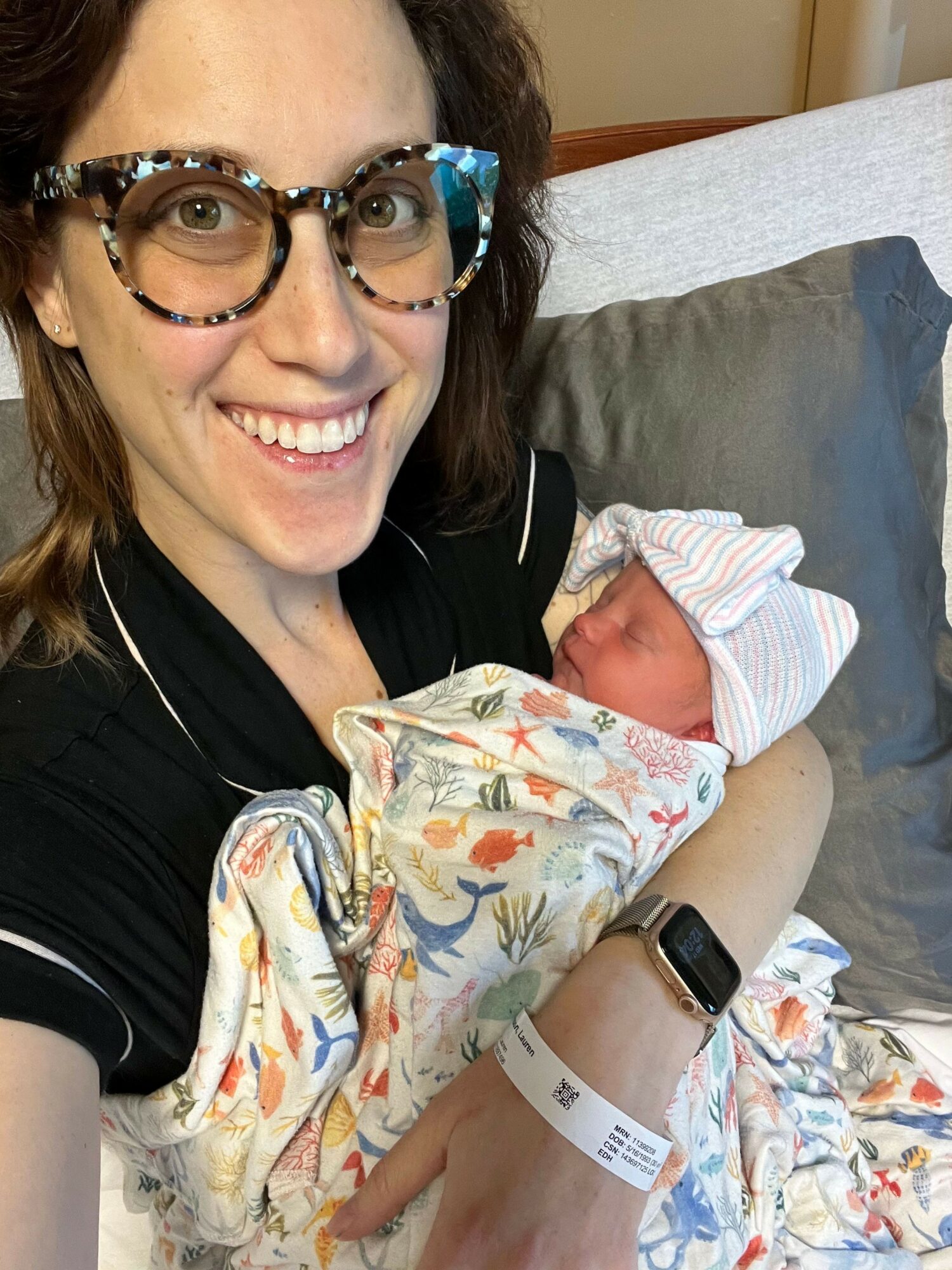 Woman with glasses smiling, holding a newborn baby wrapped in a colorful blanket, in a hospital bed.