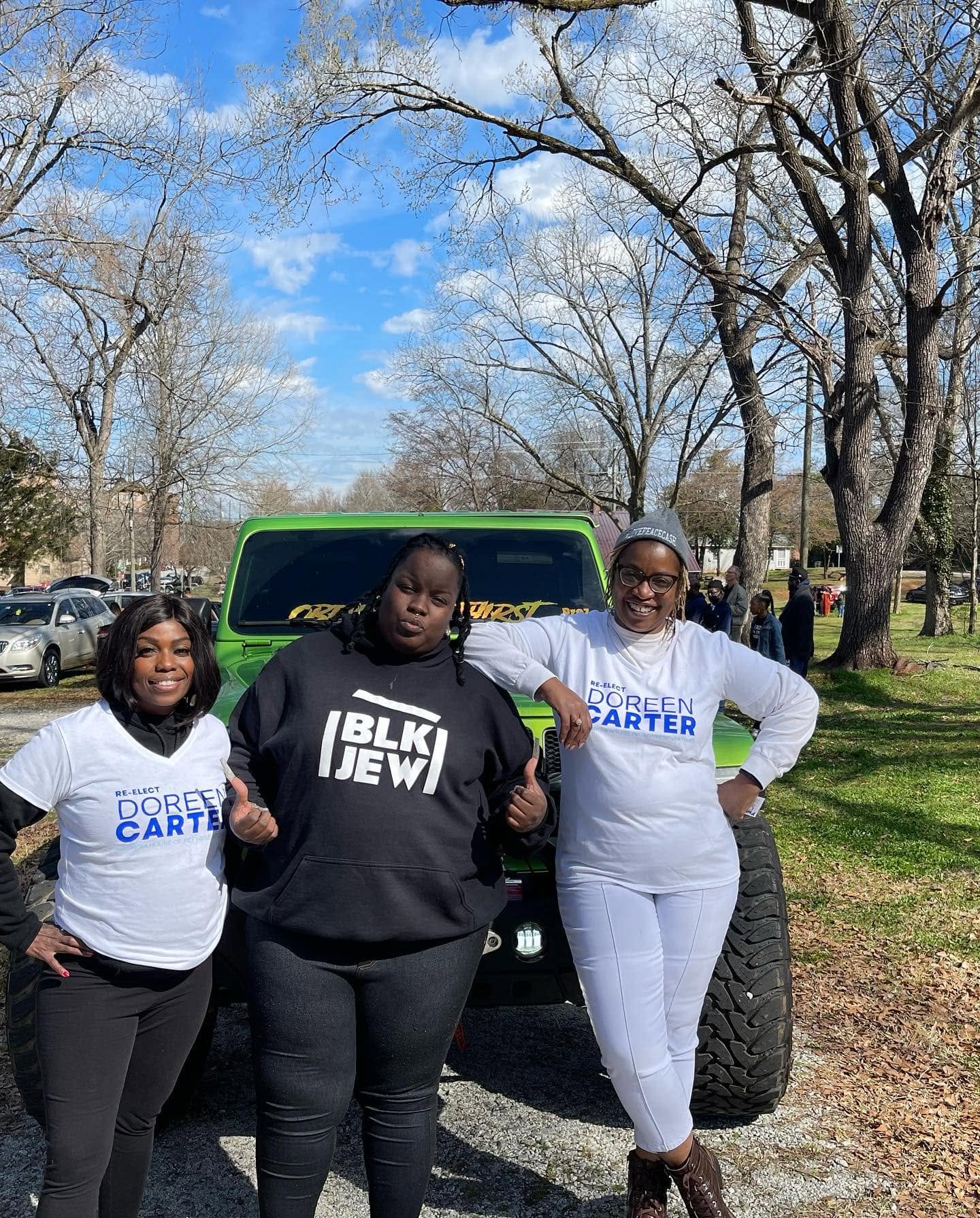 Three women standing outdoors in front of a green vehicle, smiling, with trees and a blue sky in the background.