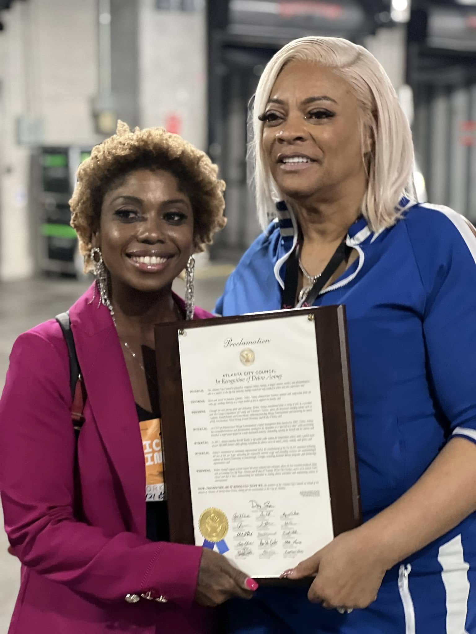 Two women holding a framed certificate, smiling, in an indoor setting with industrial background.